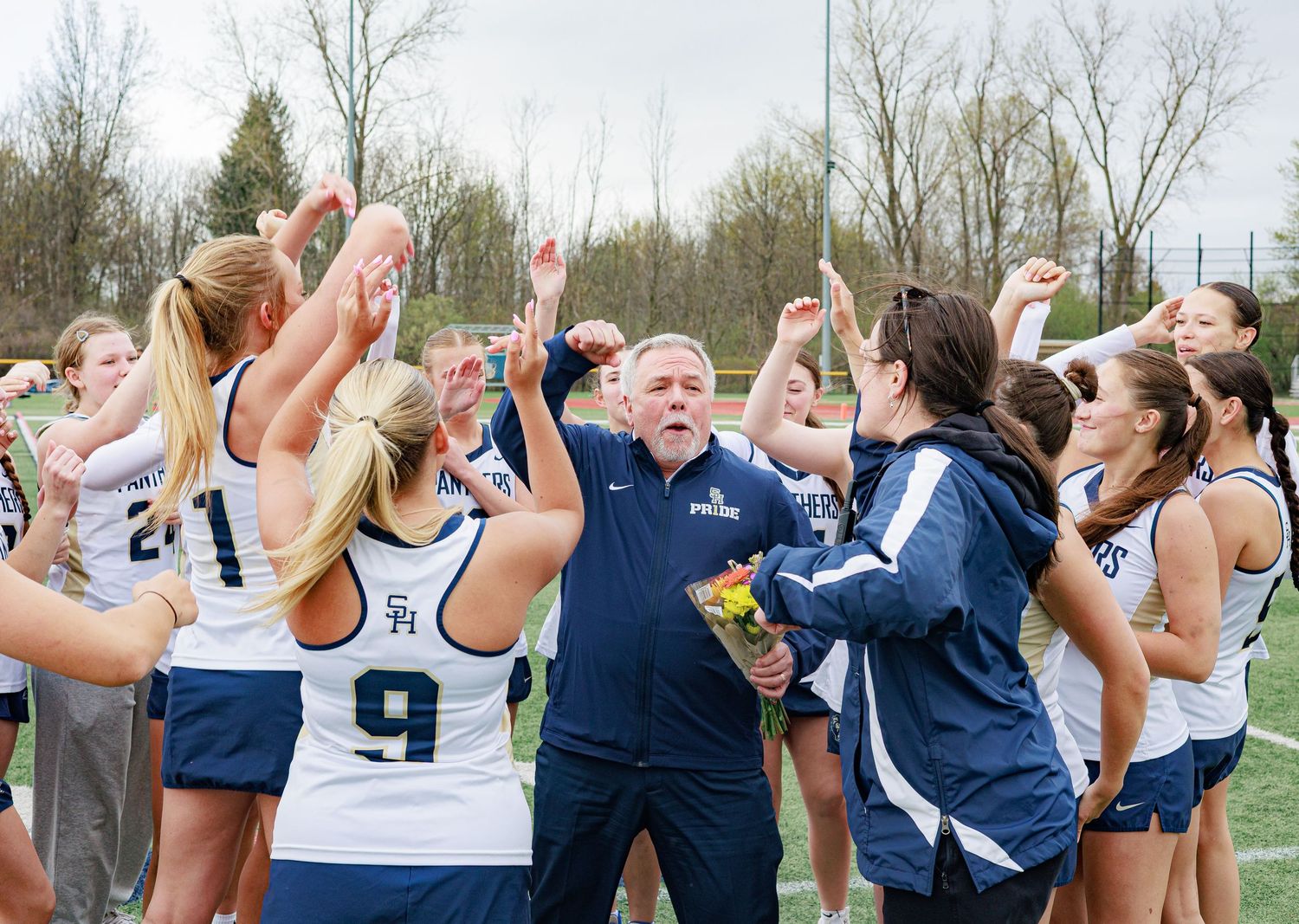 A lacrosse coach celebrates with his team of female players outdoors on a field after a game.