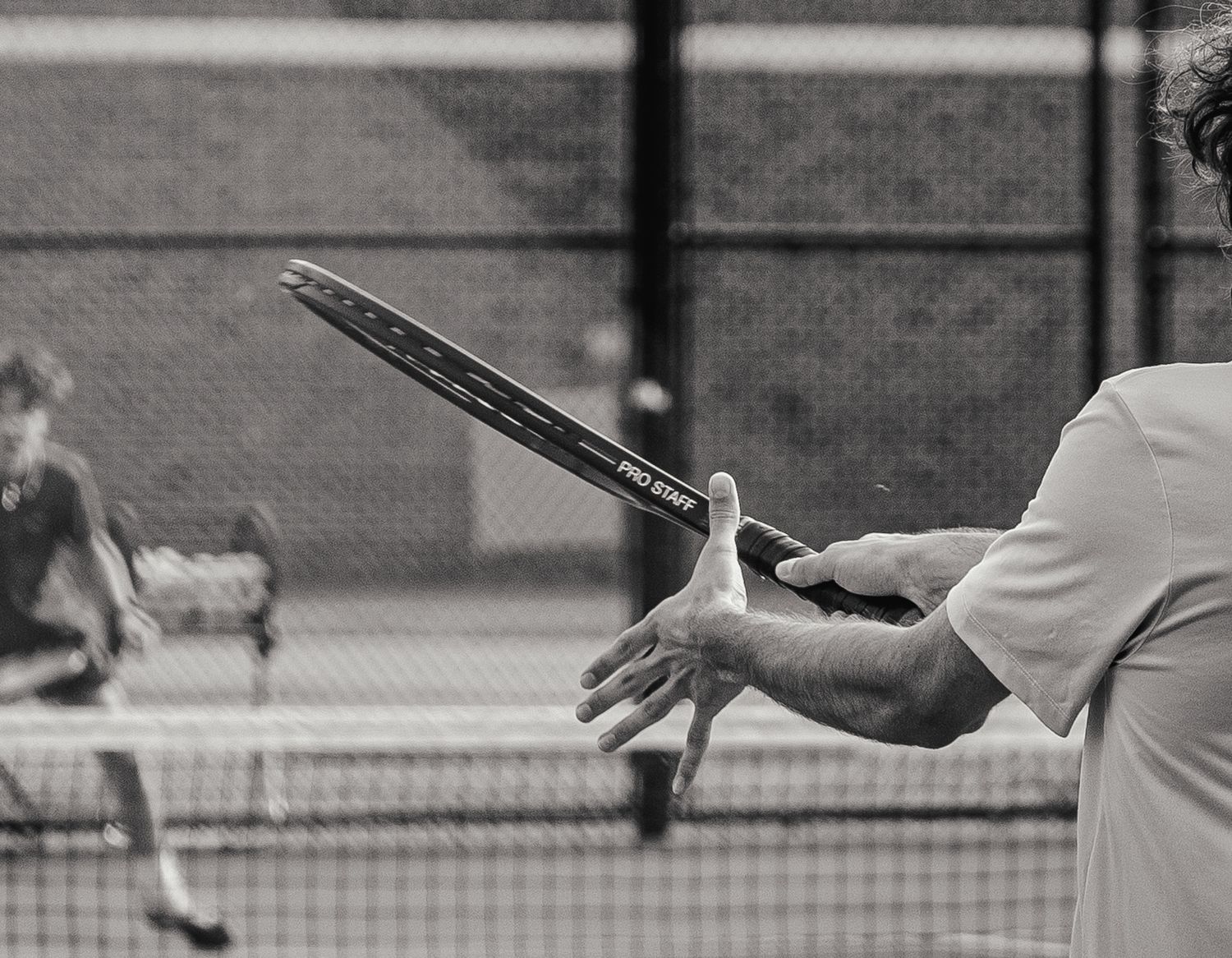 Black and white photo of a man holding a tennis racket mid-swing on an outdoor court with a net in the background.