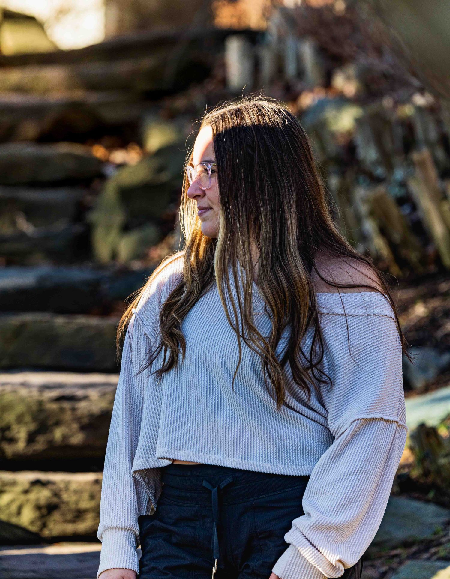 Young woman with long brown hair wearing a white ribbed crop top and dark jeans, sitting outdoors near stone steps.