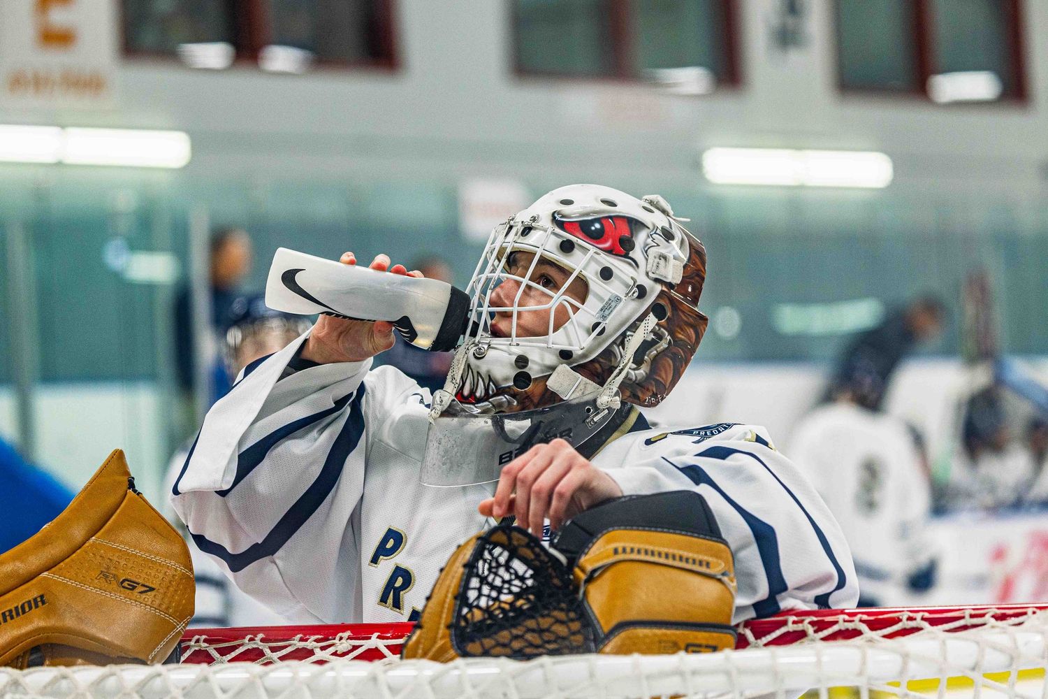 Ice hockey goalie in white uniform drinks water while resting against the net during a game.