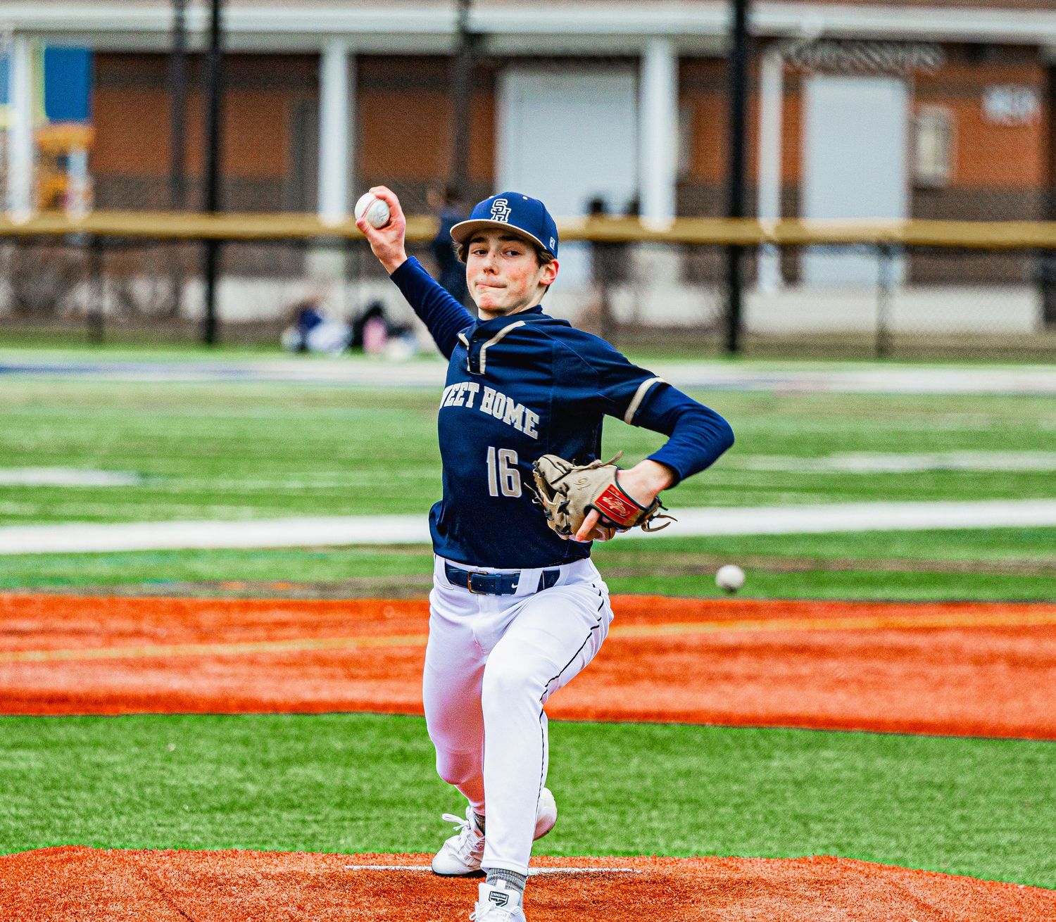 Youth baseball player wearing number 16 jersey pitching from the mound on an orange artificial turf field.