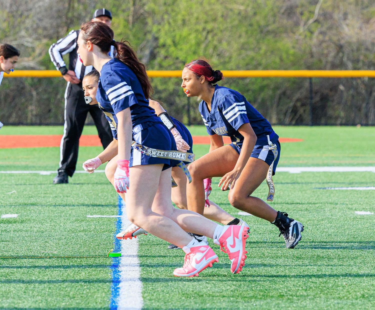 Female flag football players in blue uniforms line up at scrimmage line on turf field during a game.