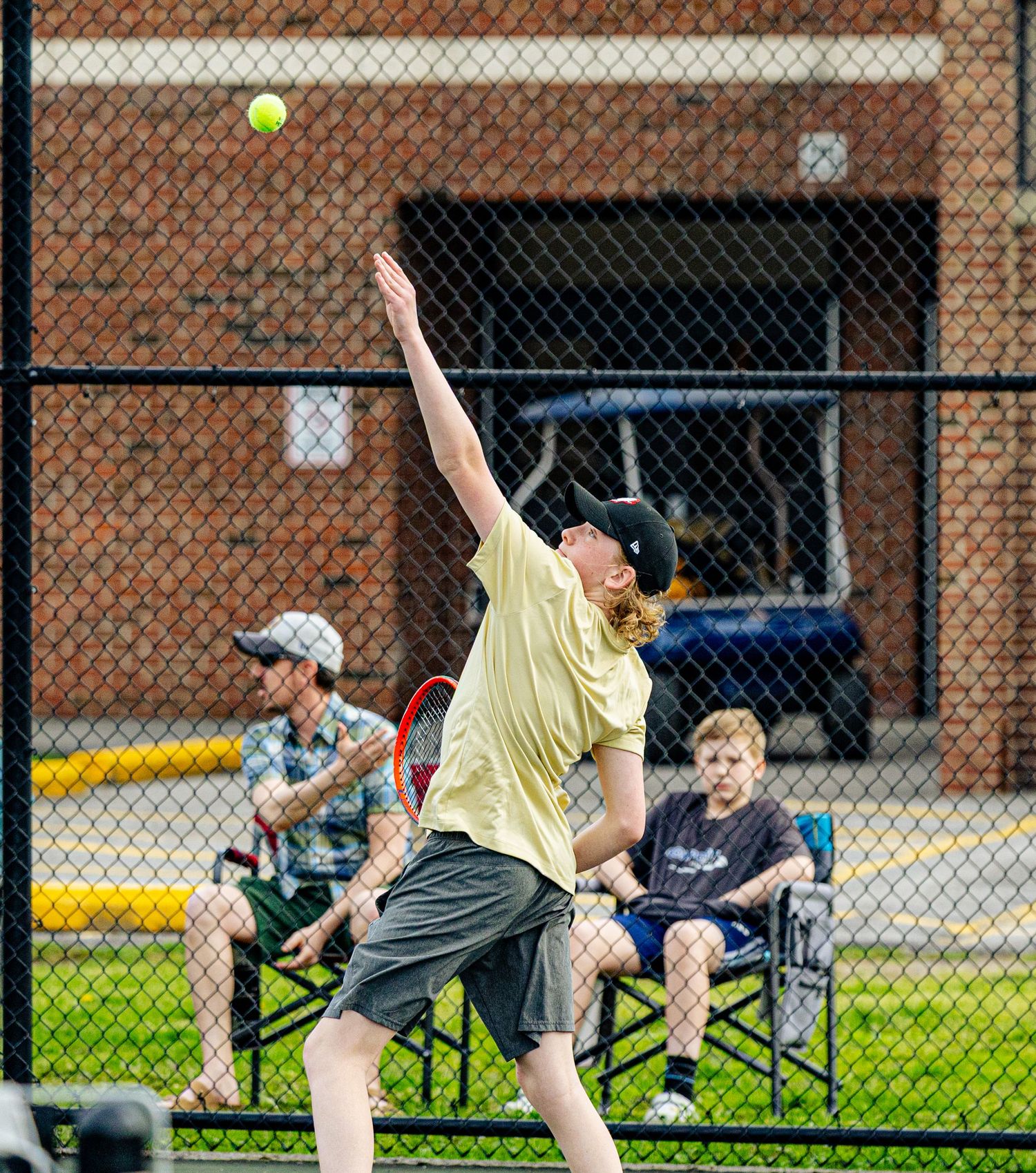 A young tennis player serves the ball on an outdoor court while spectators watch from chairs in the background.