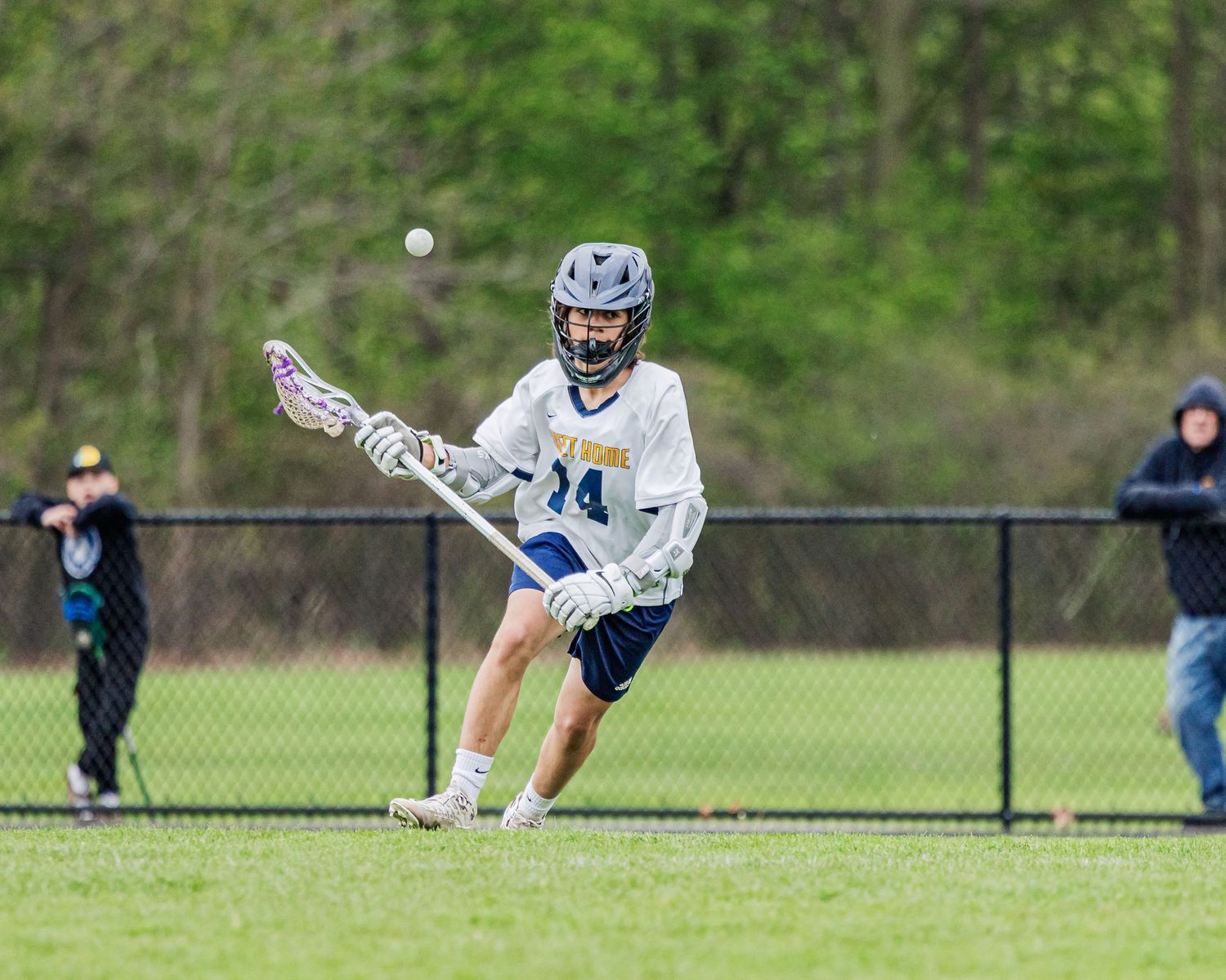 Youth lacrosse player in blue and white uniform scooping ball during outdoor game on green field.