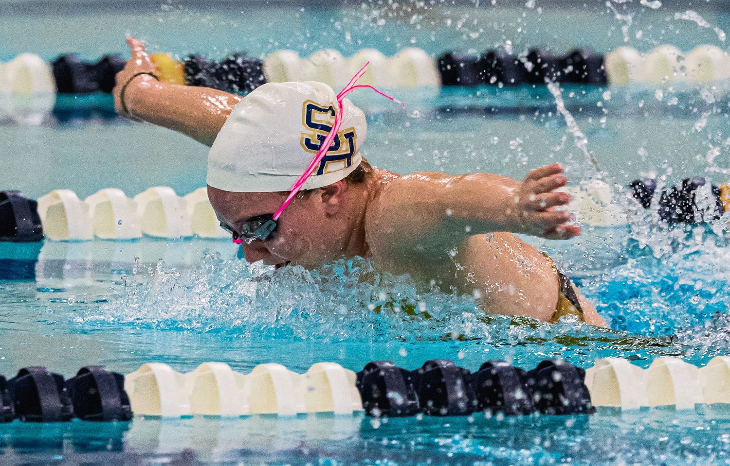 Competitive swimmer performing butterfly stroke in pool, wearing white cap and goggles during a swim meet.