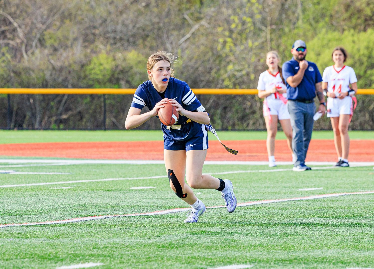 Female flag football quarterback in blue uniform runs with football during outdoor game on green turf field.