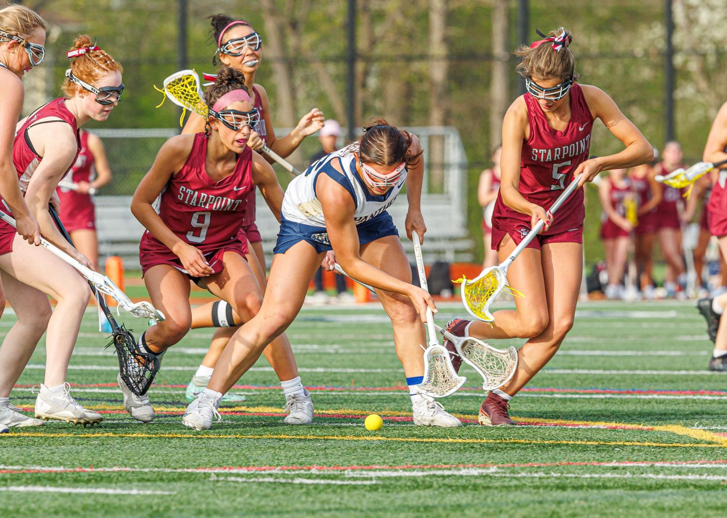 Girls lacrosse players compete for the ball during an intense match, wearing red and white uniforms on a turf field.