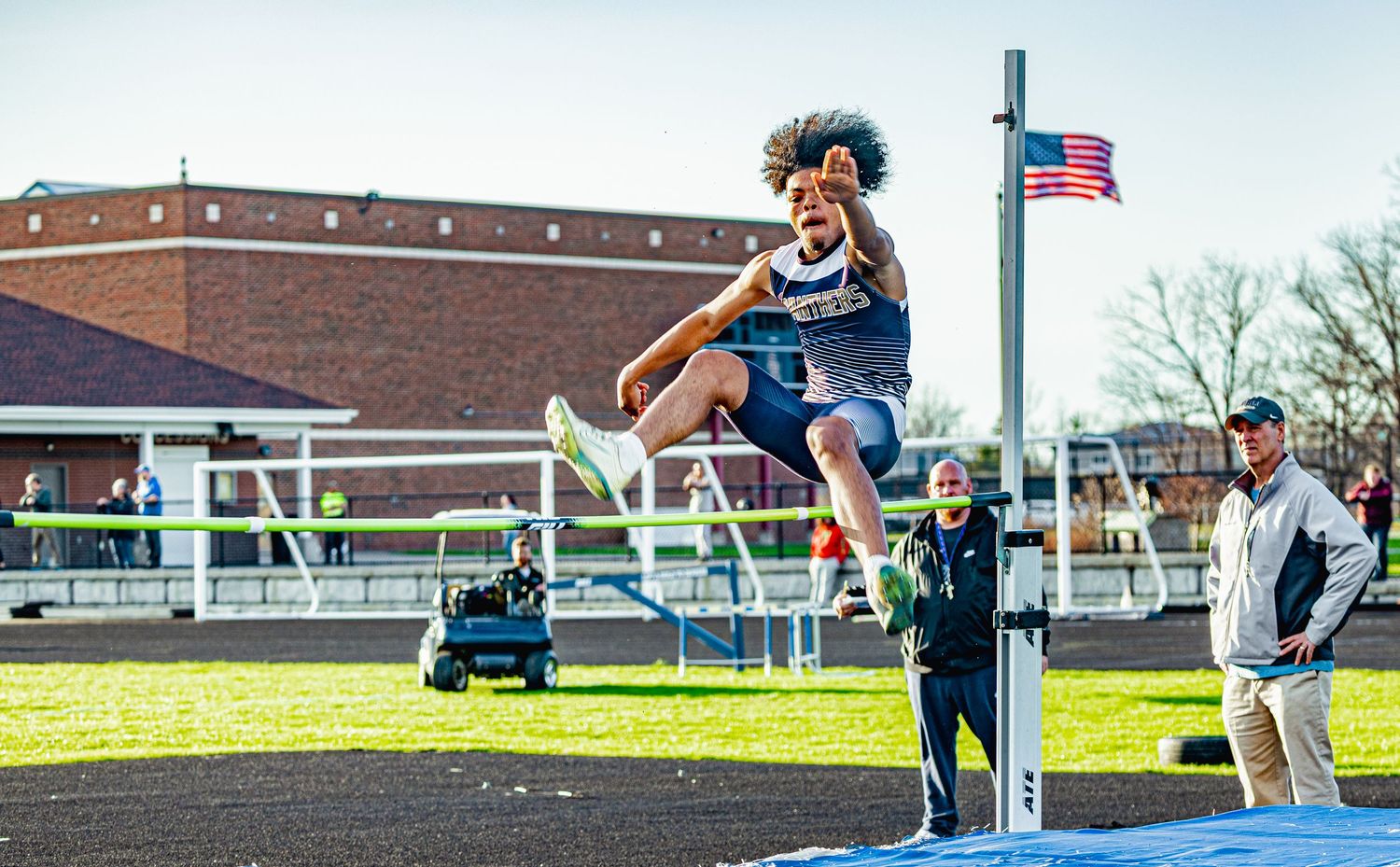 Athlete performs a high jump at an outdoor track meet with an American flag and spectators in the background.