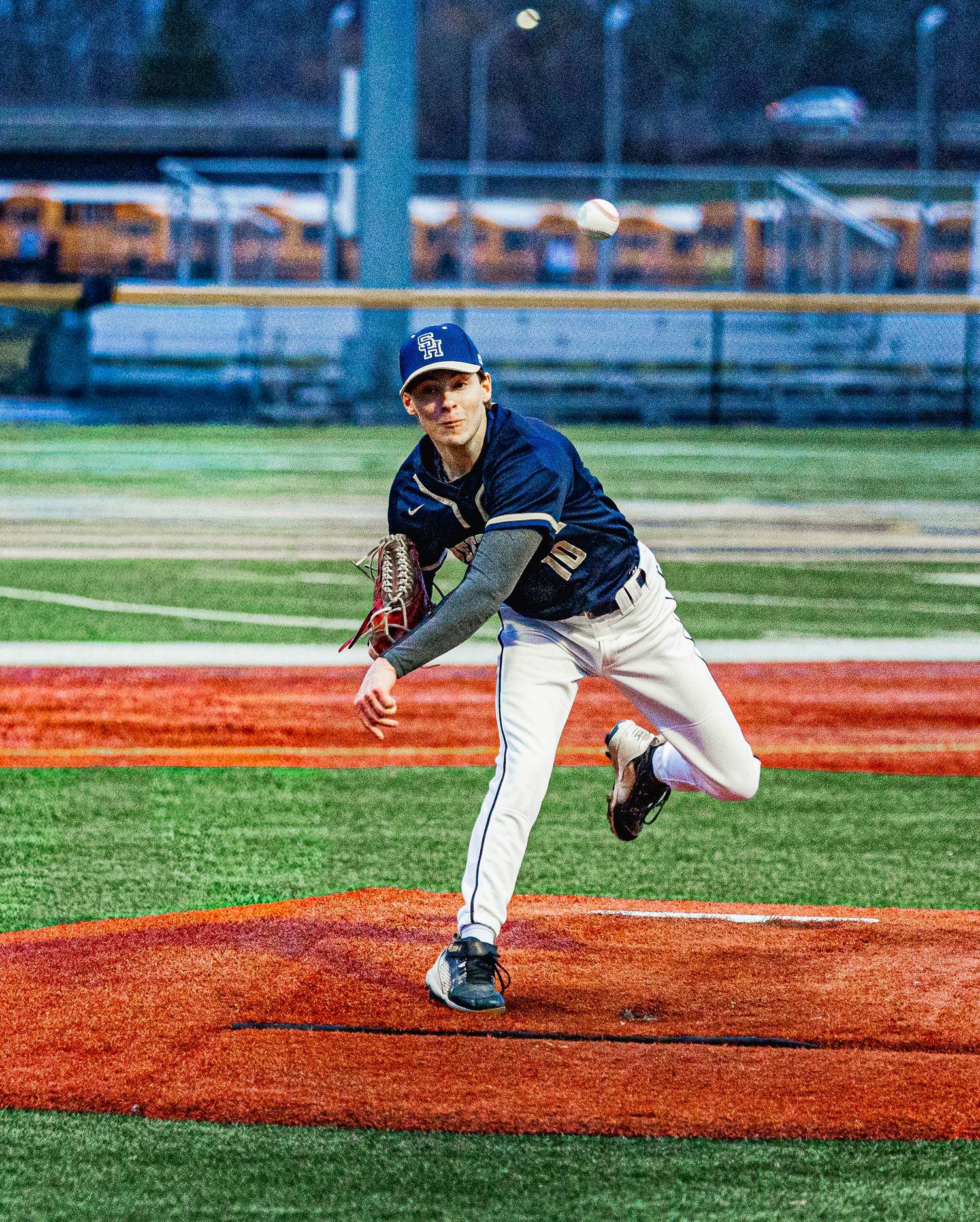 Baseball pitcher in navy uniform mid-throw on red mound during evening game.
