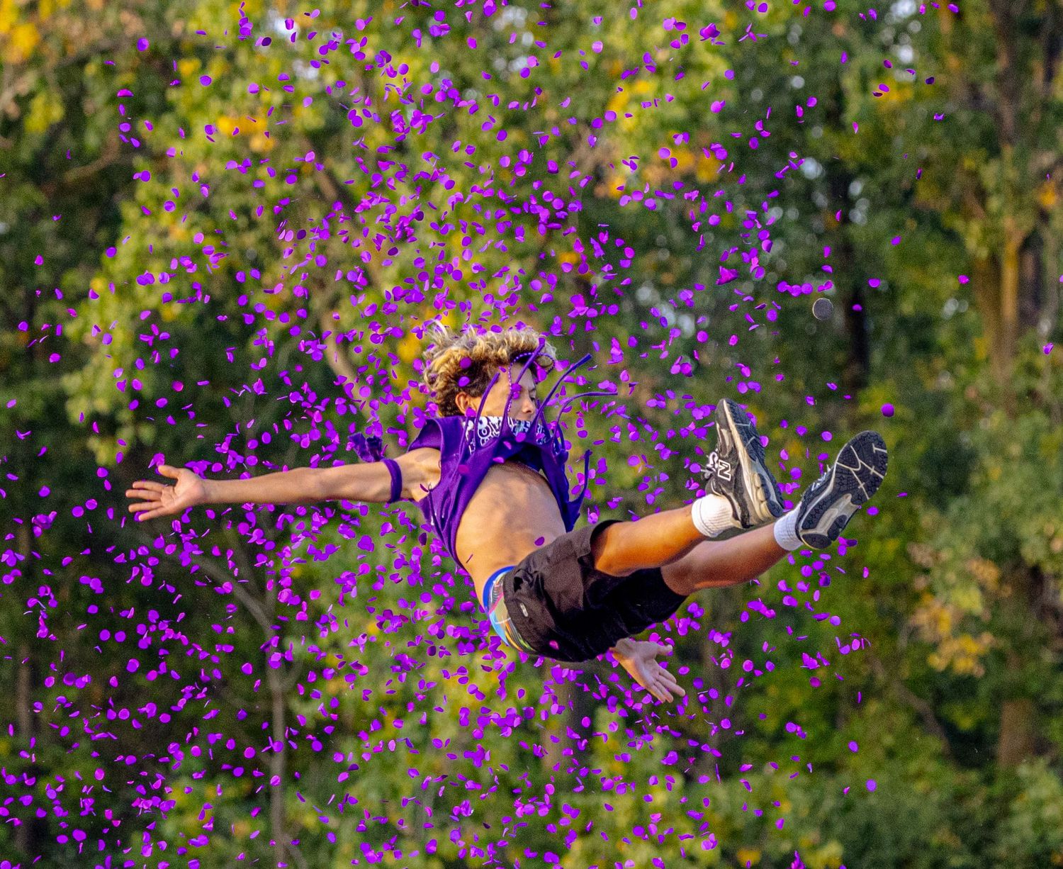 Person in purple outfit leaping mid-air surrounded by scattered purple flowers with green trees in background.