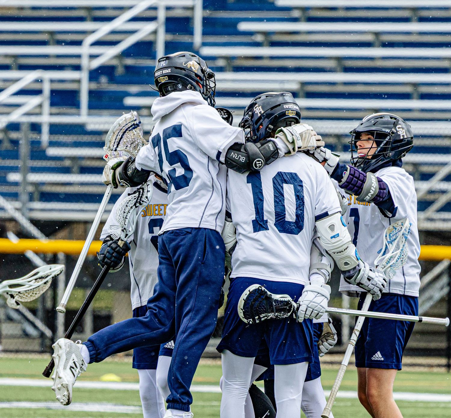 Lacrosse players in white and navy uniforms celebrating together on the field during a game.