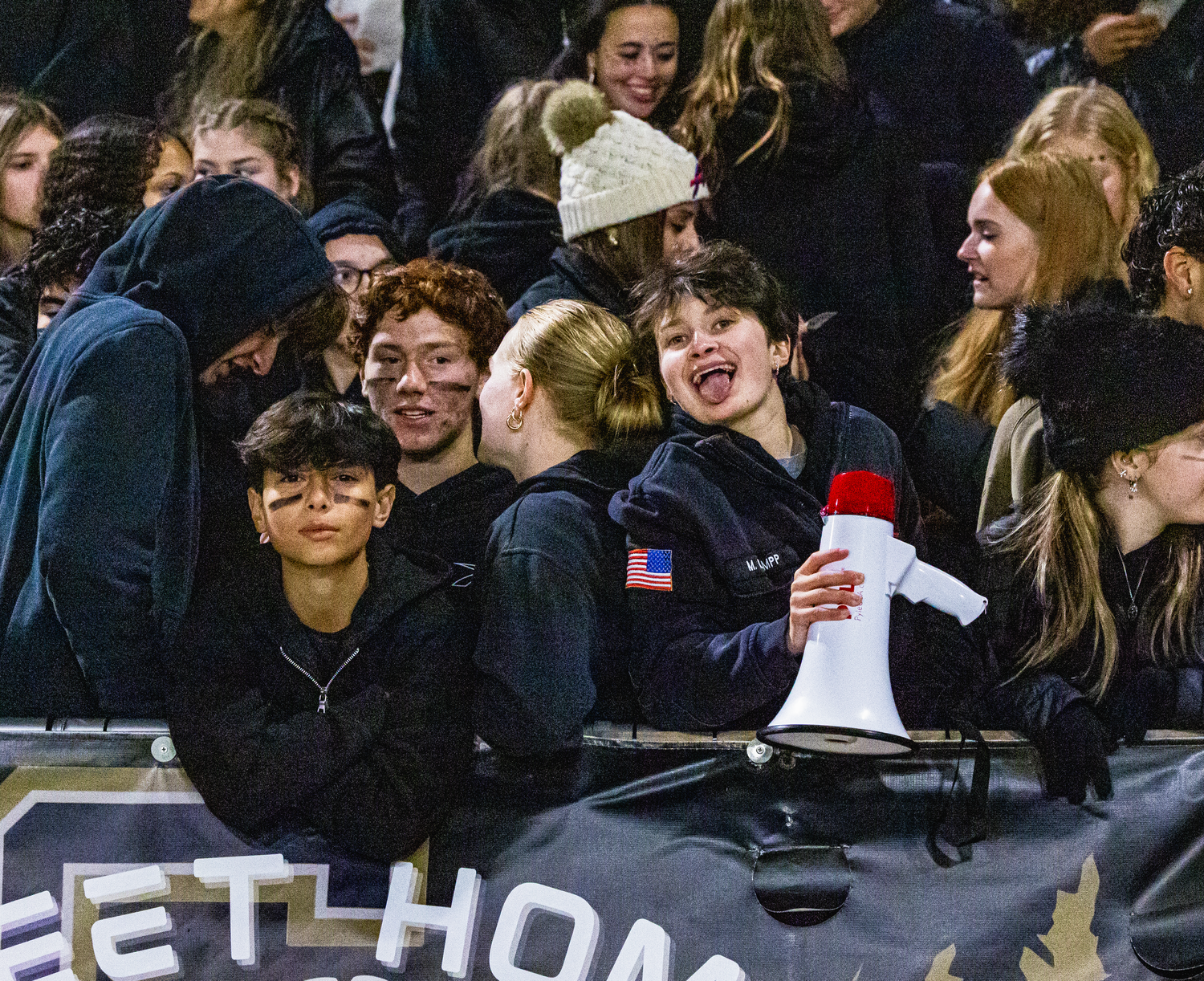 Excited students cheering in bleachers at a night football game, one holding a red and white megaphone.