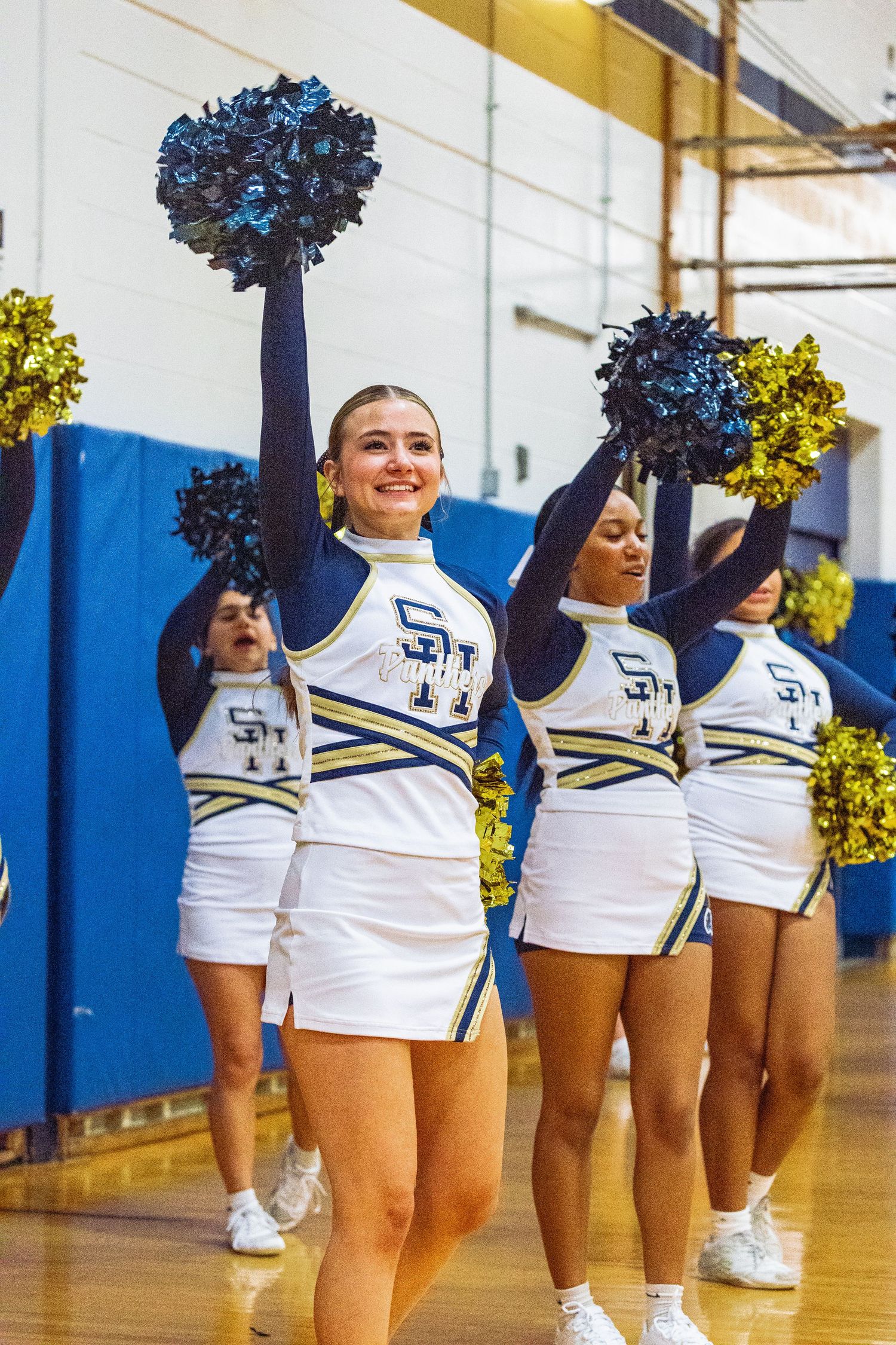 Cheerleaders in white and blue uniforms wave navy and gold pom-poms during a high school pep rally in a gymnasium.