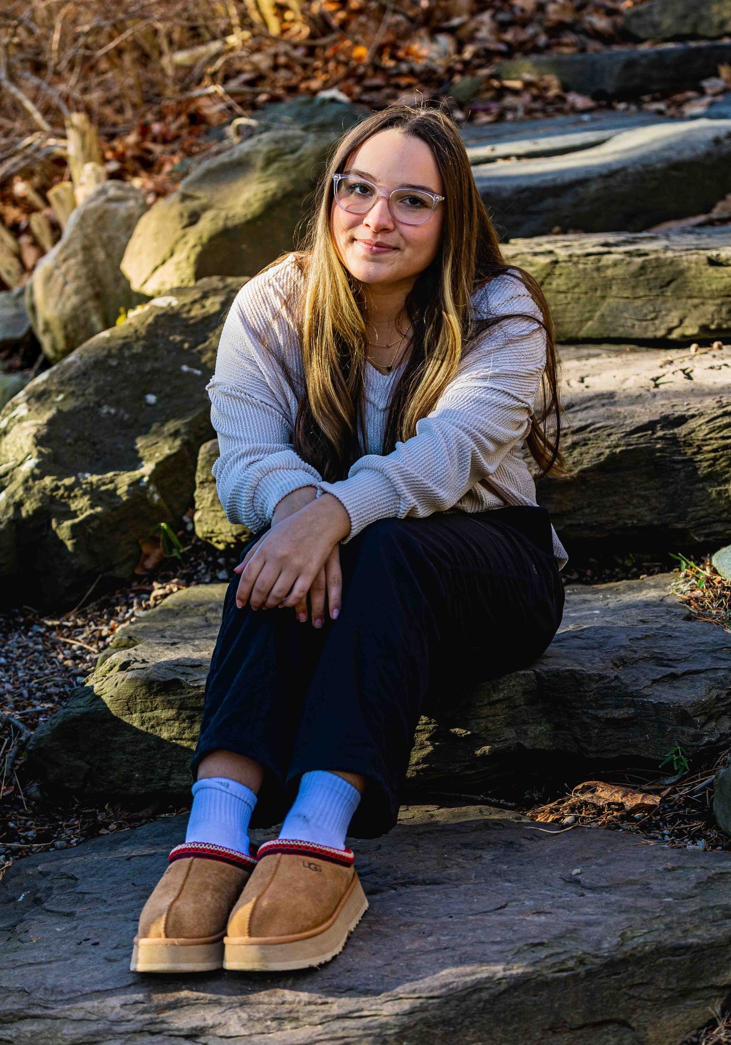 Young woman with glasses sits on rocky steps outdoors, wearing a cream sweater, dark pants, and tan UGG slippers.