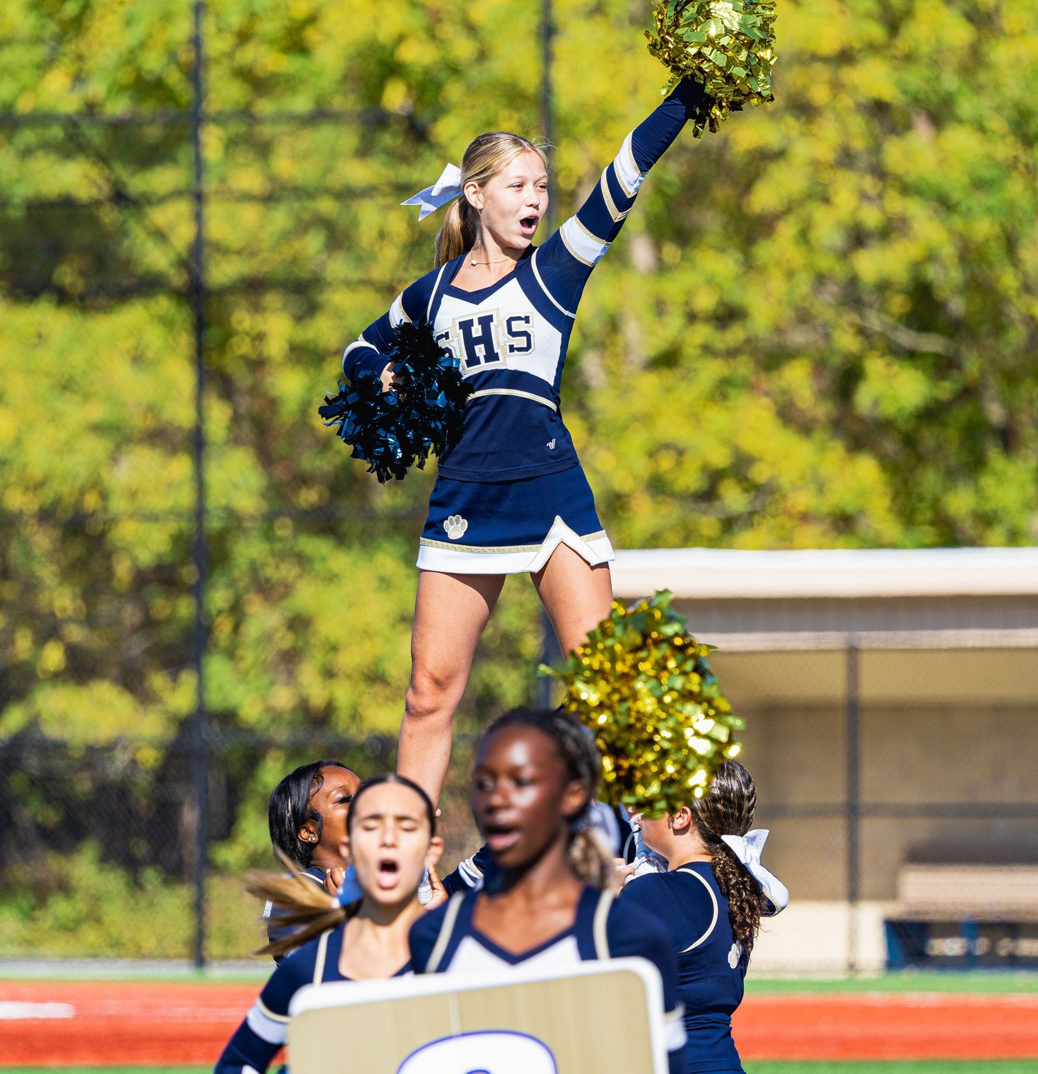 Cheerleader elevated by teammates performs a stunt with pom-poms raised at an outdoor high school football game.
