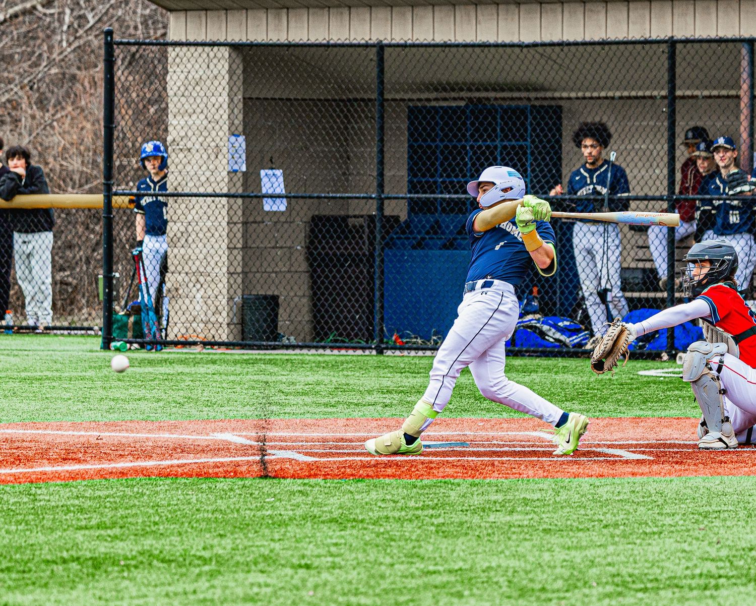 Baseball batter swinging at pitch during a youth league game on a green field with teammates watching from the dugout.