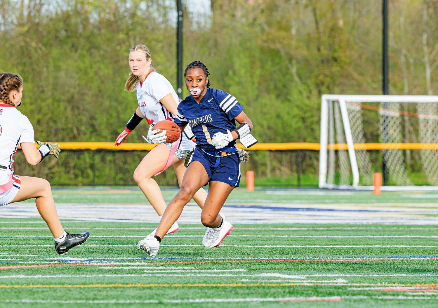 Female flag football player in navy uniform runs with ball past opponents on turf.