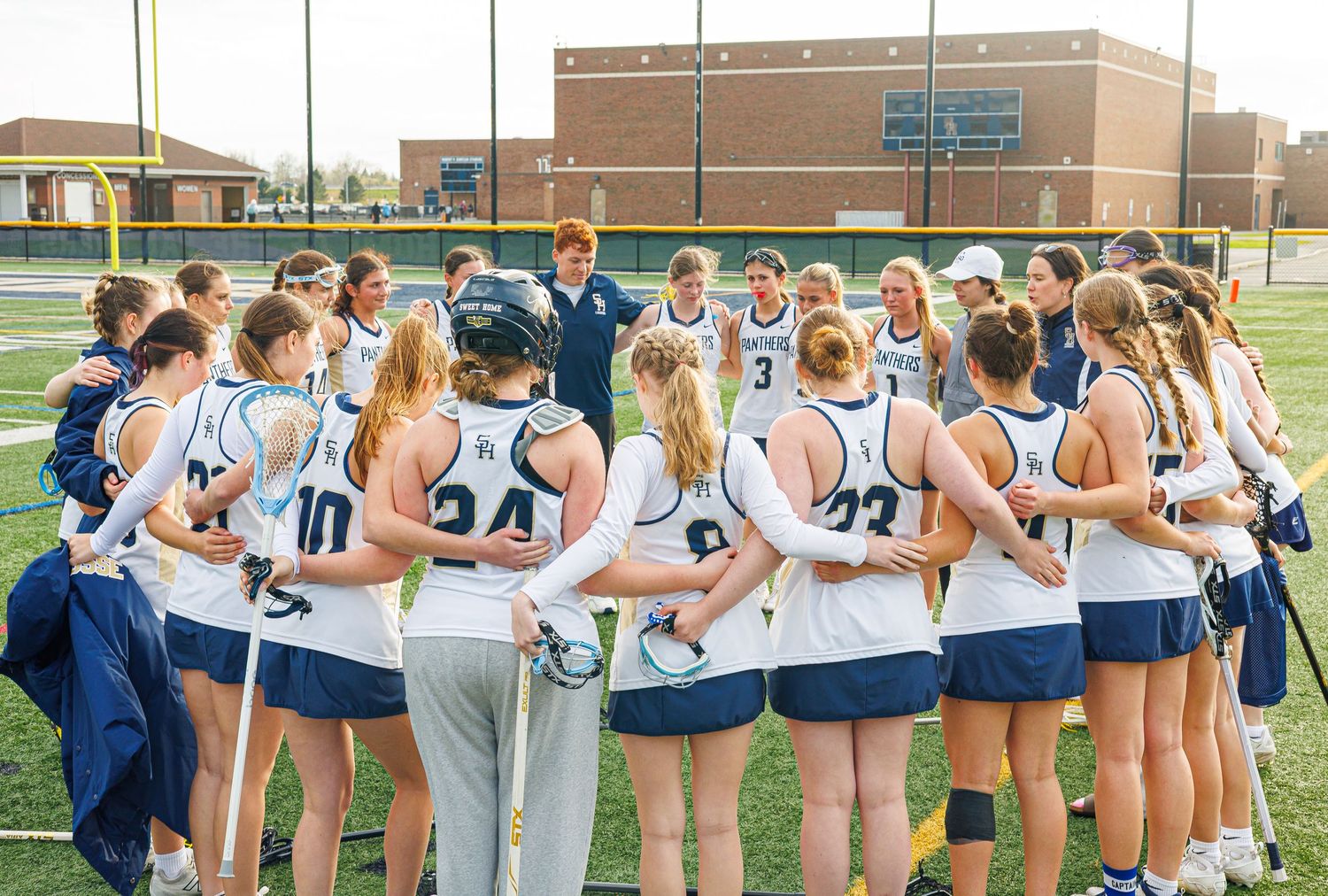 Girls lacrosse team huddles together with coach on turf field after a game.