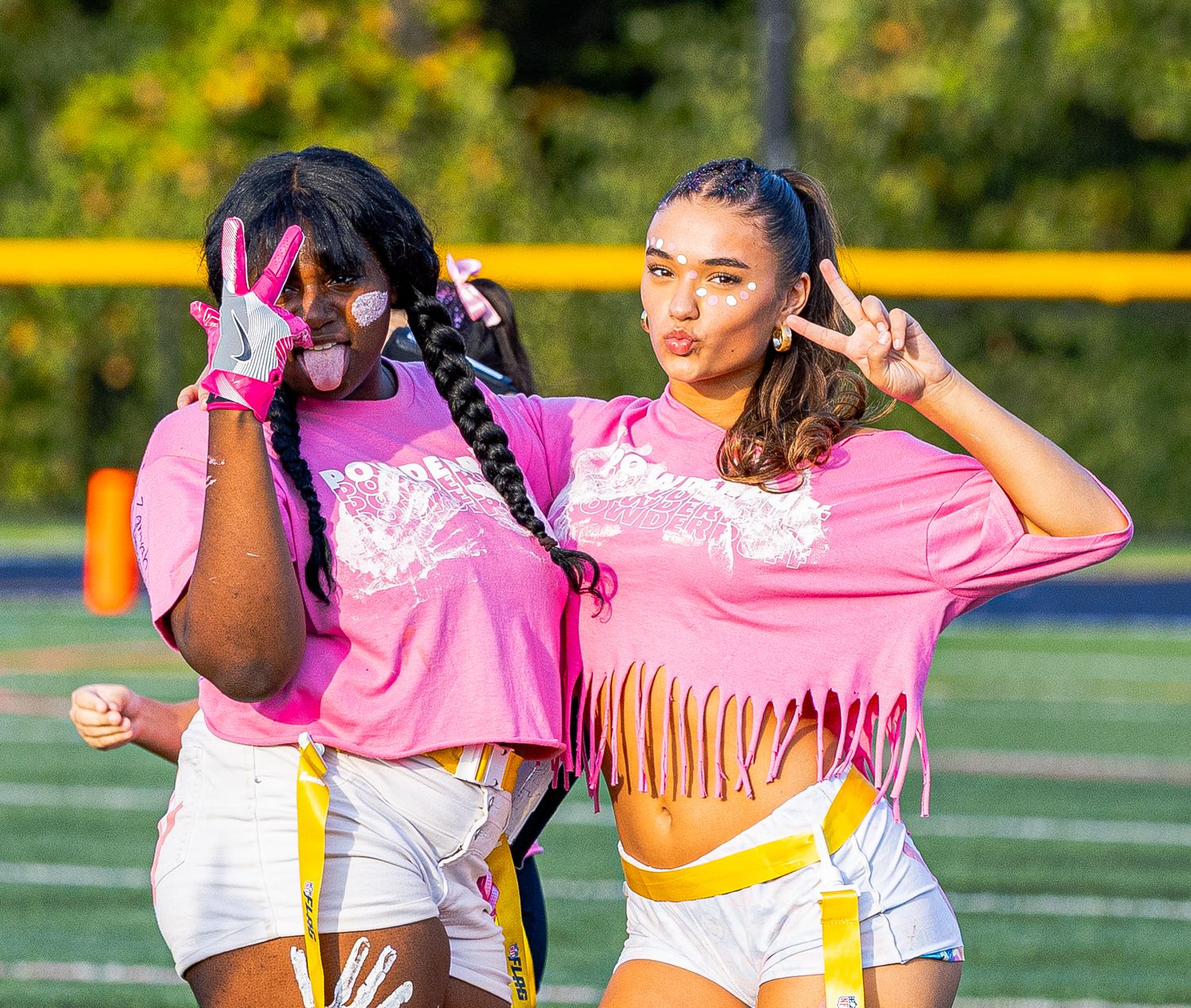 Two girls in matching pink fringe tops and white shorts pose with peace signs on a football field.