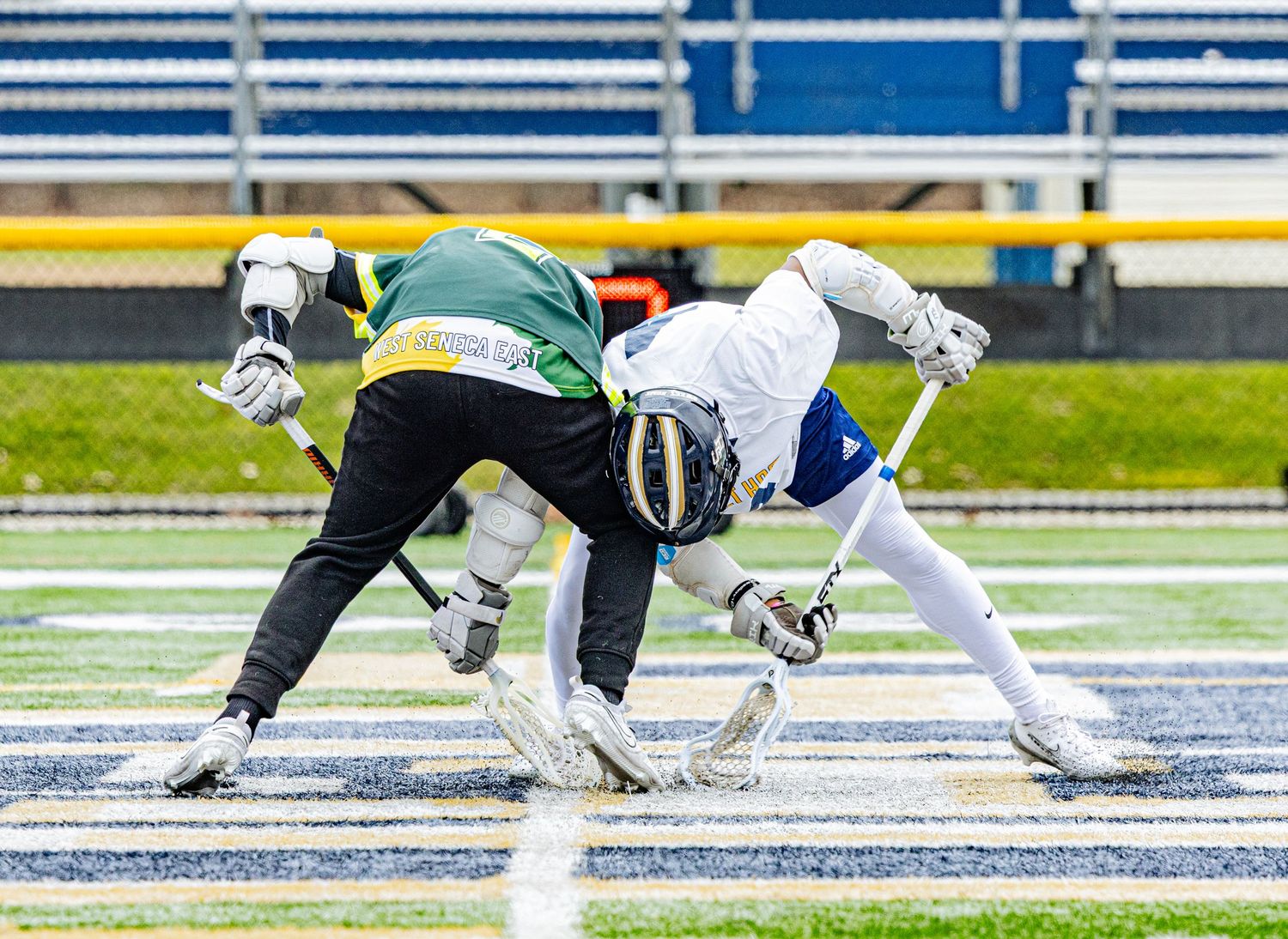 Two lacrosse players compete for the ball during a face-off on a blue turf field in a stadium.