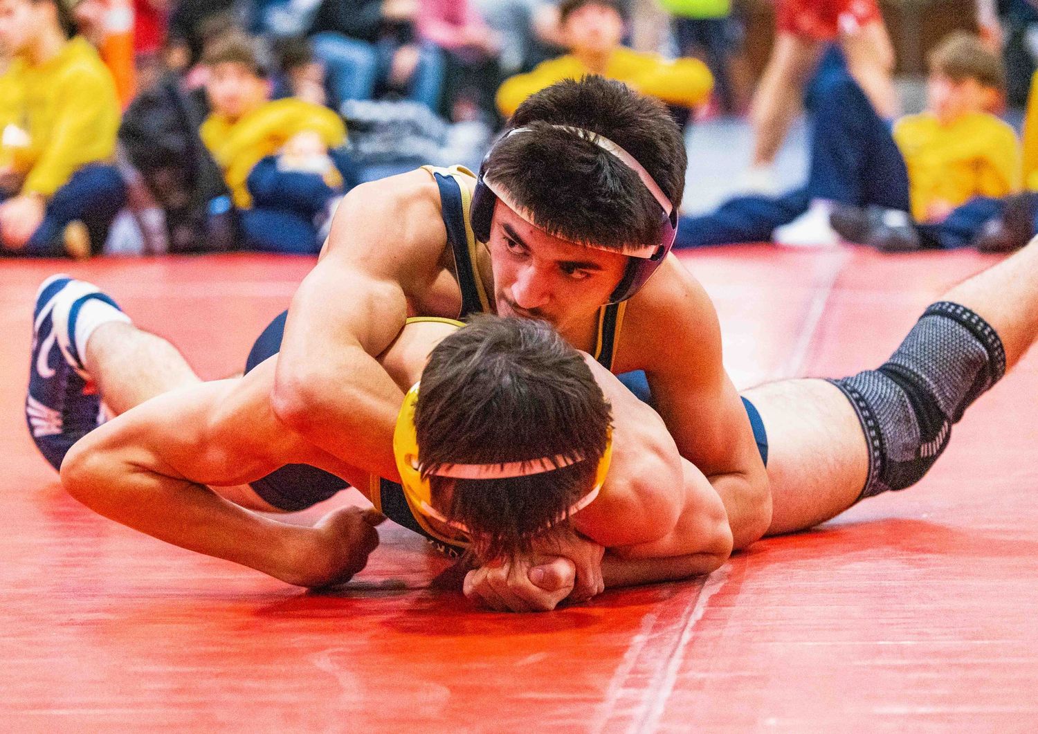 Two wrestlers compete on a red mat, with one athlete pinning his opponent down during an intense wrestling match.