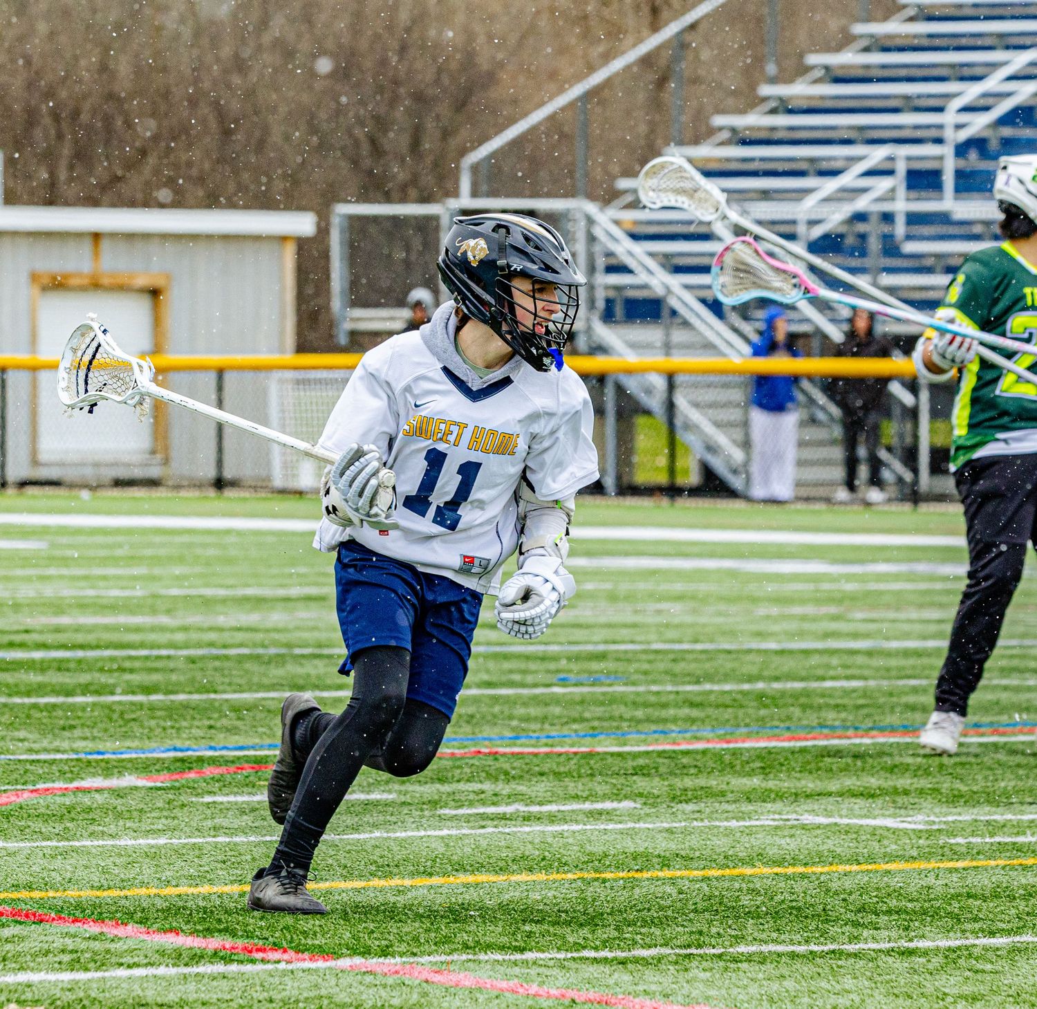 Player #11 in white lacrosse uniform sprints down field during outdoor game.
