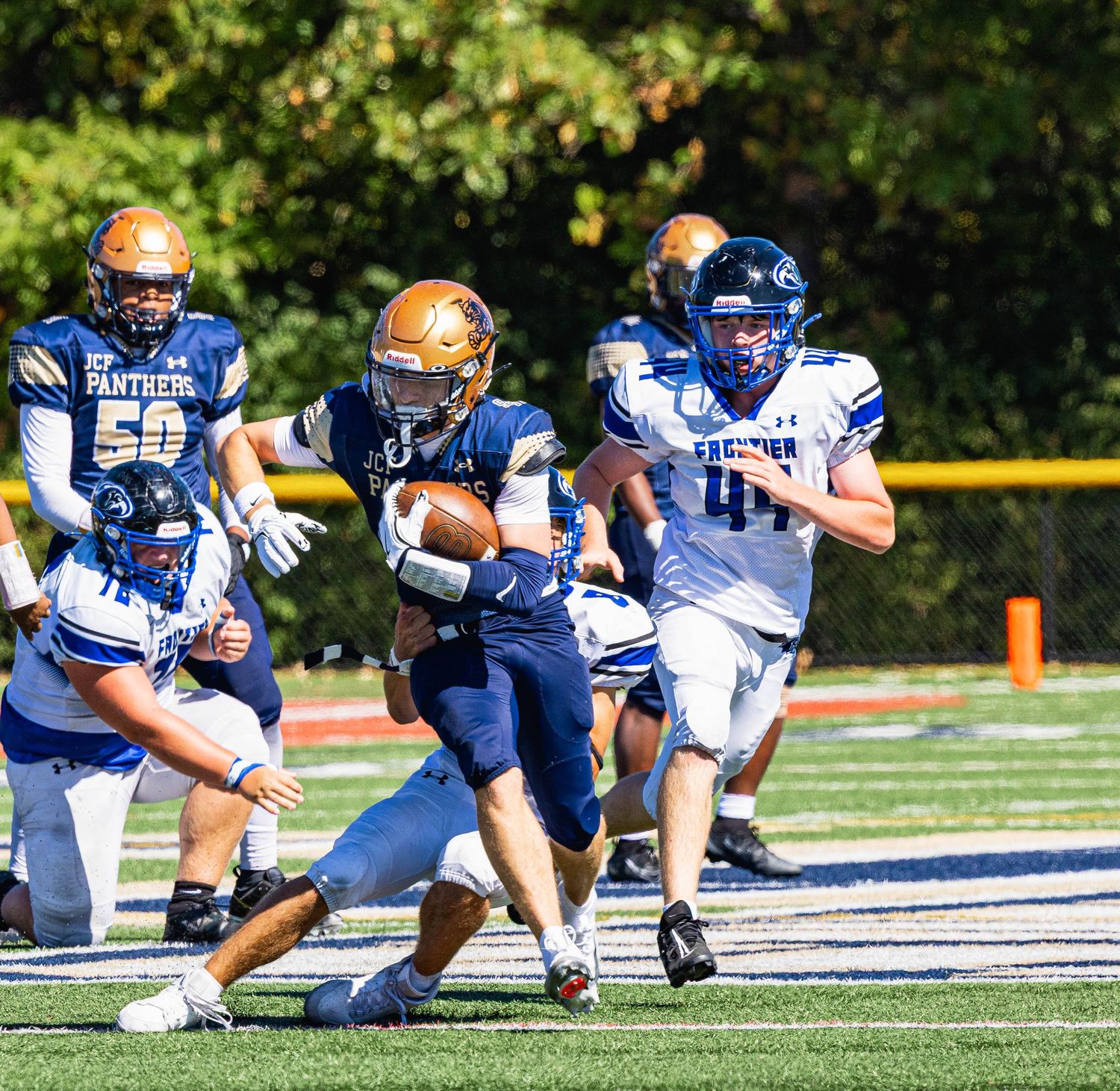 Panthers football player runs with the ball during a sunny outdoor game as opposing team players in blue uniforms give chase.