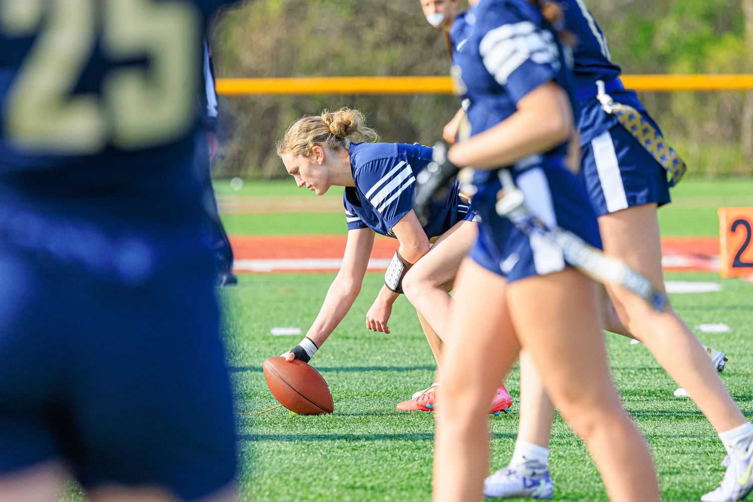 Girl in navy uniform picks up football during flag football game on turf field.