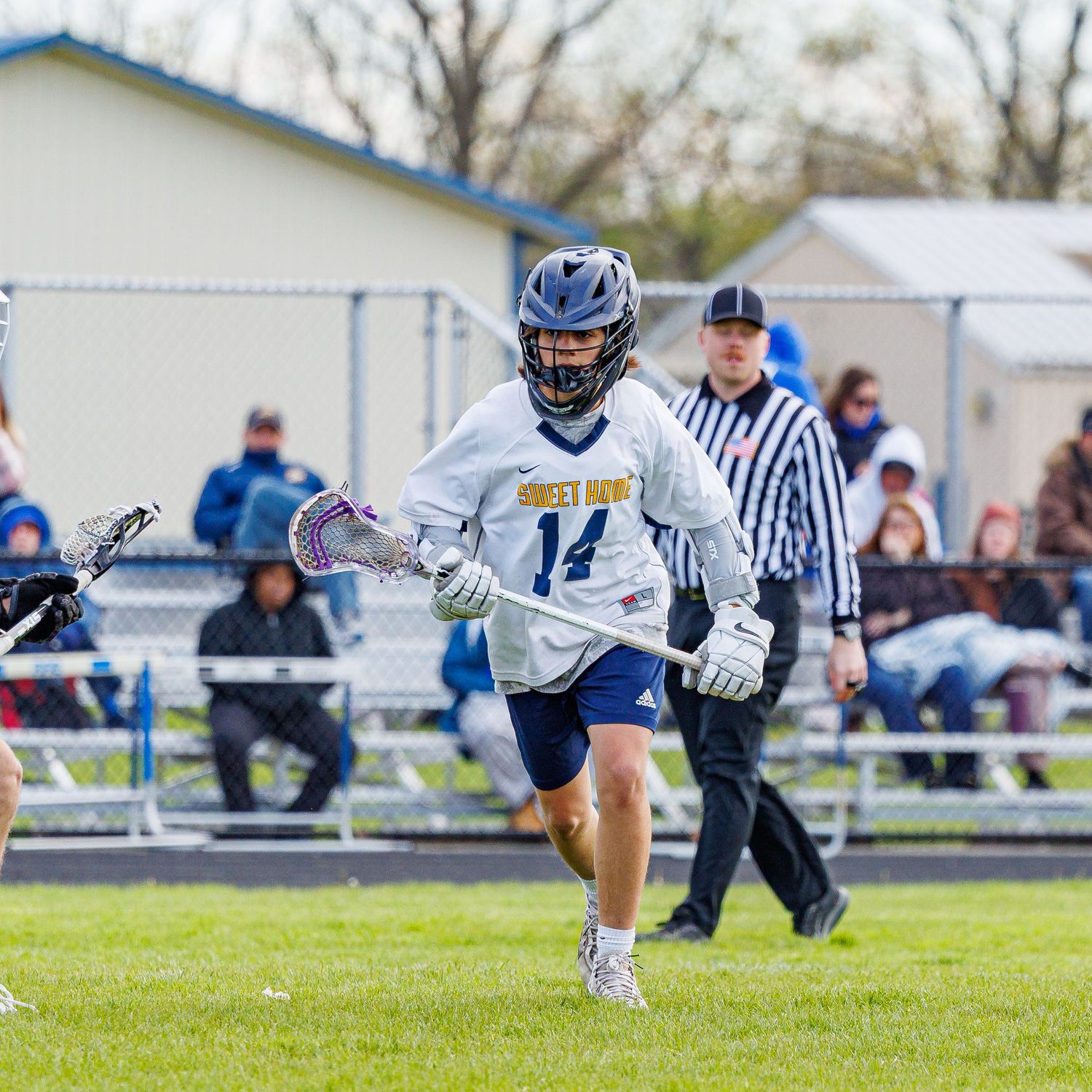 Youth lacrosse player #14 in blue and white uniform runs with stick during outdoor game with referee in background.
