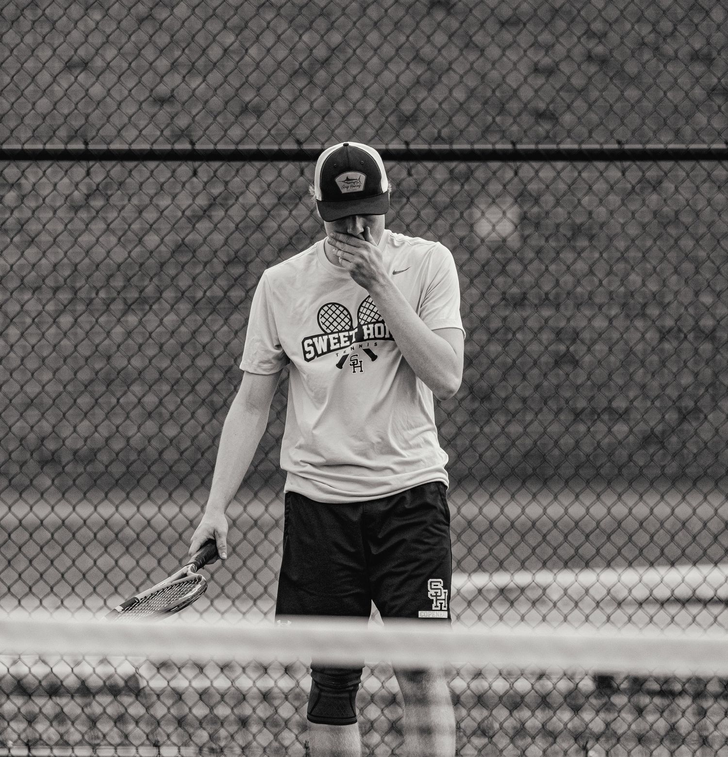 Black and white photo of a male tennis player in a cap and Nike shirt holding a racket on an outdoor court.