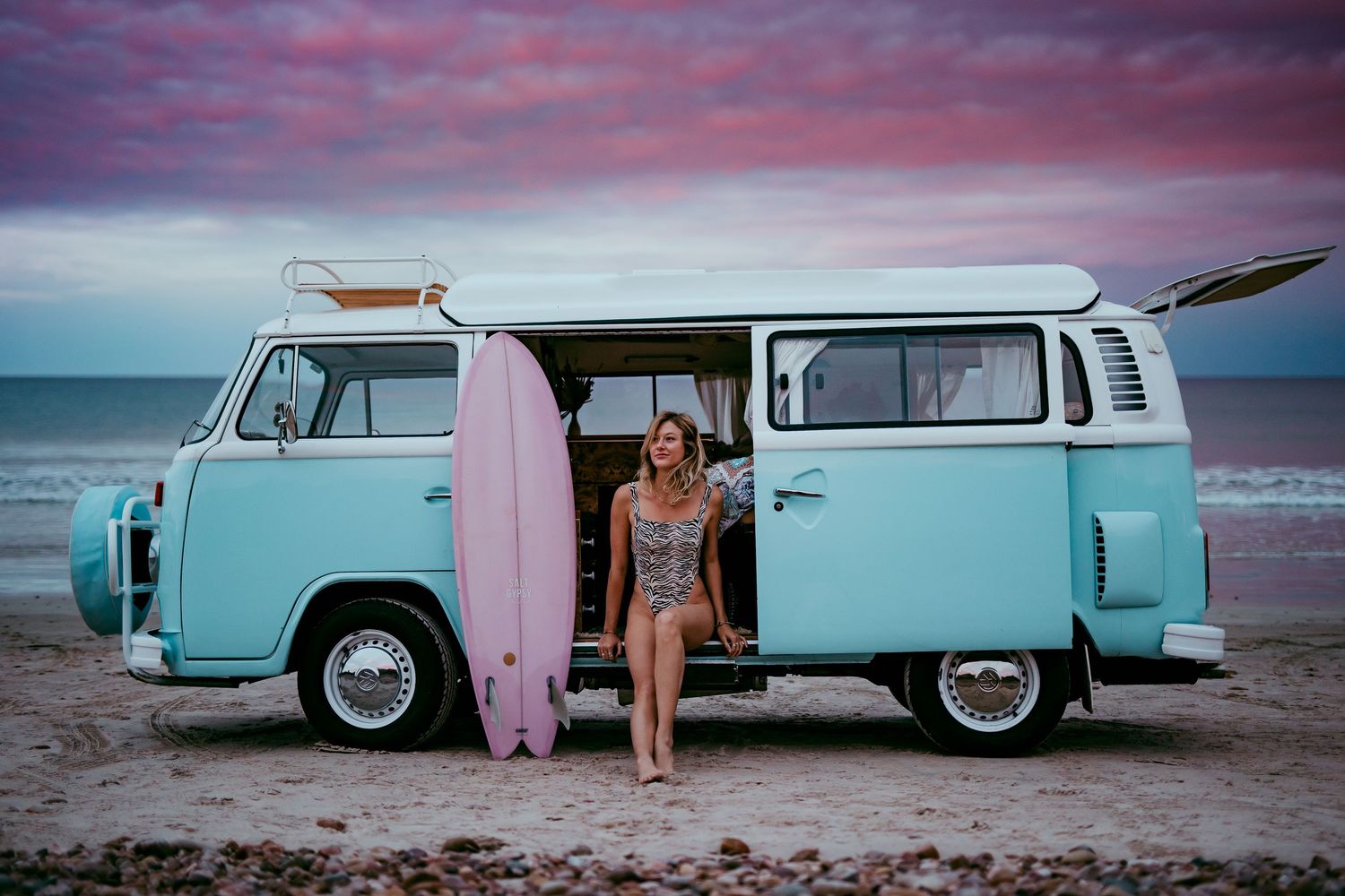 Woman sitting in a vintage Kombi van with a surfboard beside her, under a pastel pink and blue fairy floss sky at sunset