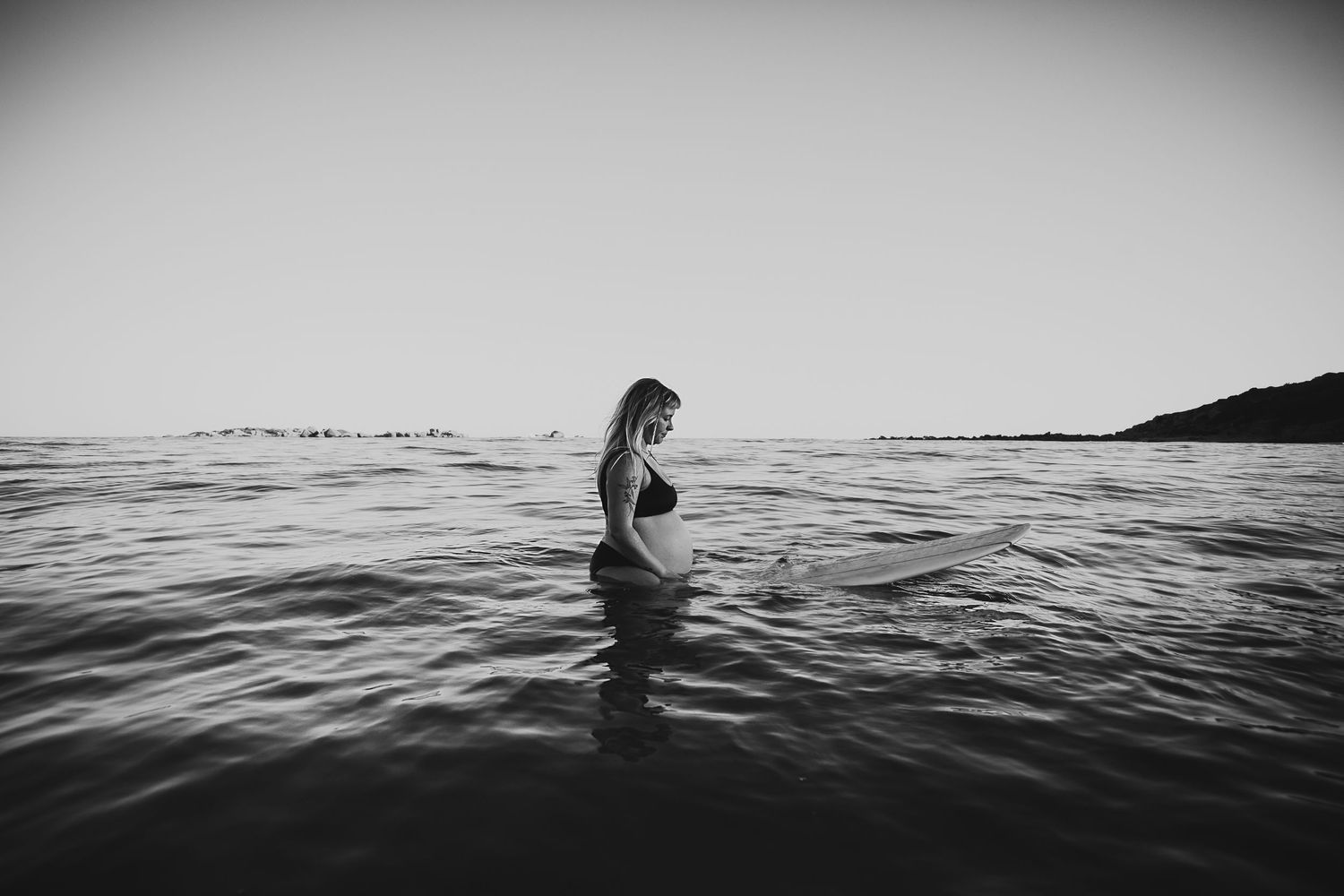 Pregnant lady sitting on a surfboard in the calm ocean. Adelaide maternity and underwater photographer - Raised by Water Photography