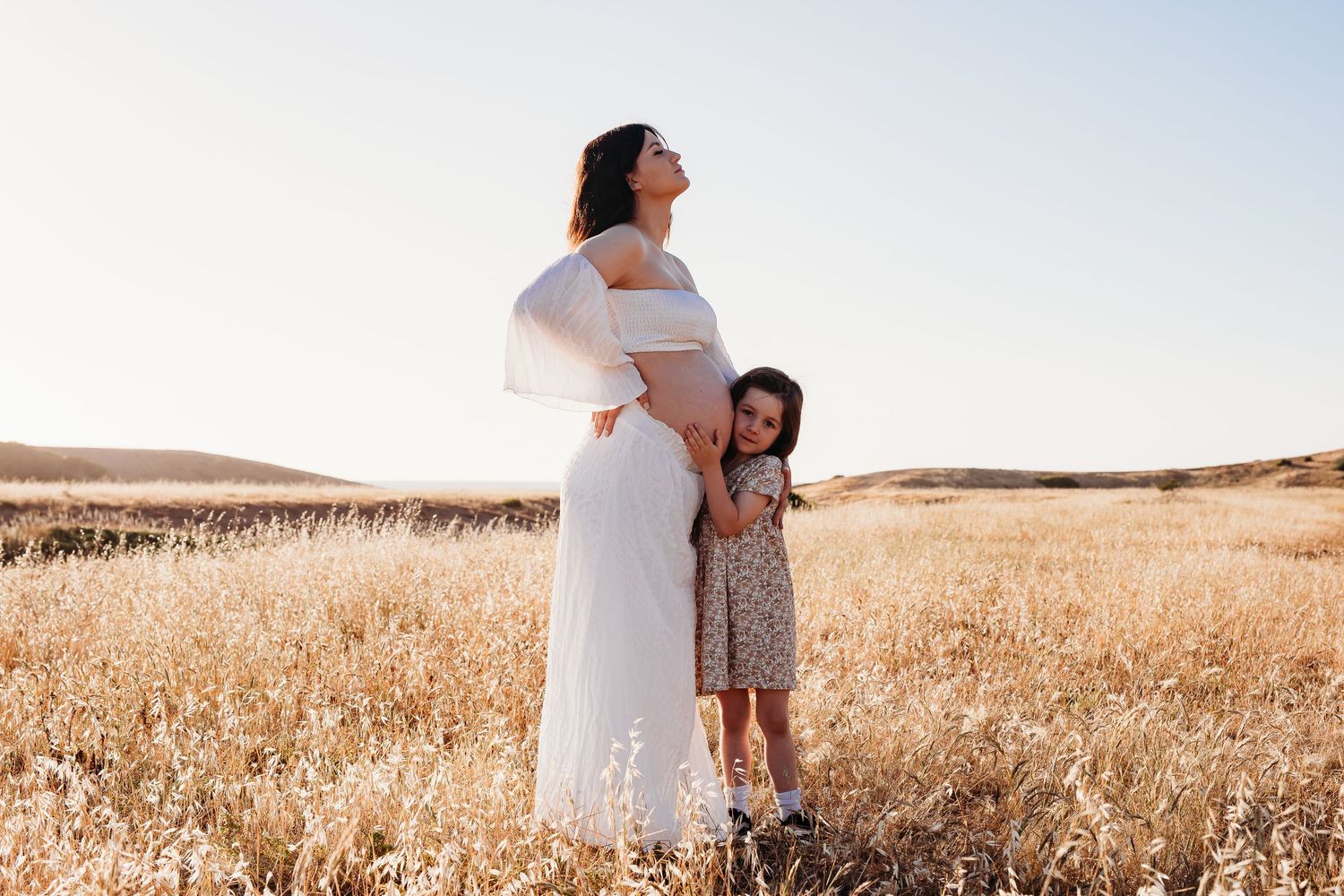 Tender moment of young girl with her ear to mother’s baby bump in golden grass – South Australia family photography