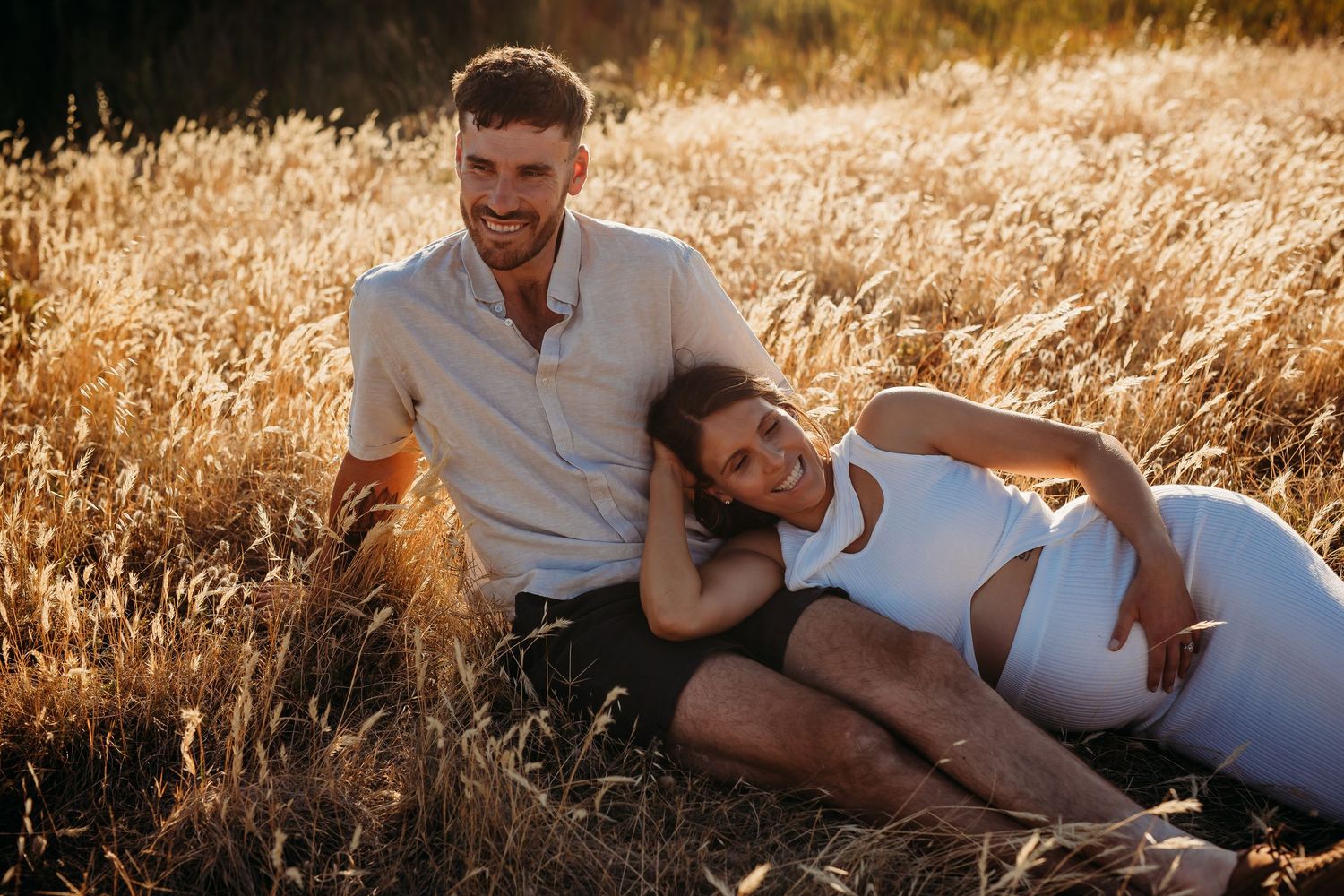 Pregnant mum lying comfortably on partner’s legs in sunlit golden field – Raised by Water Photography, Adelaide