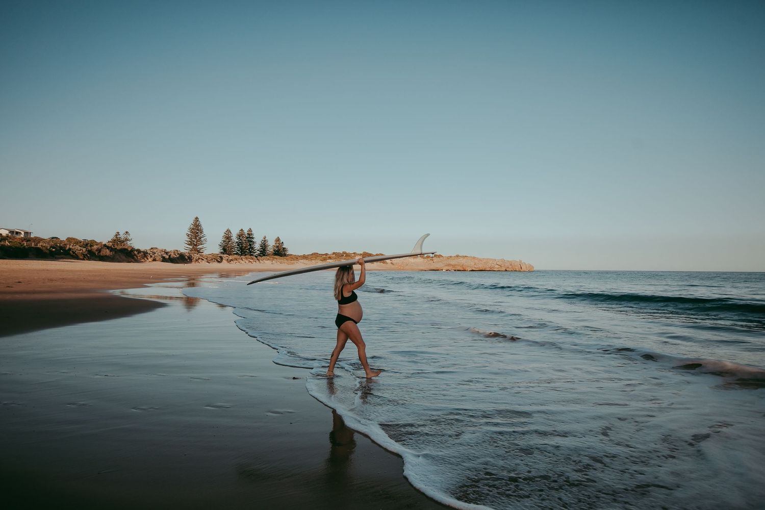 Pregnant woman carrying surfboard on head walking into ocean at Adelaide beach – Raised by Water Photography