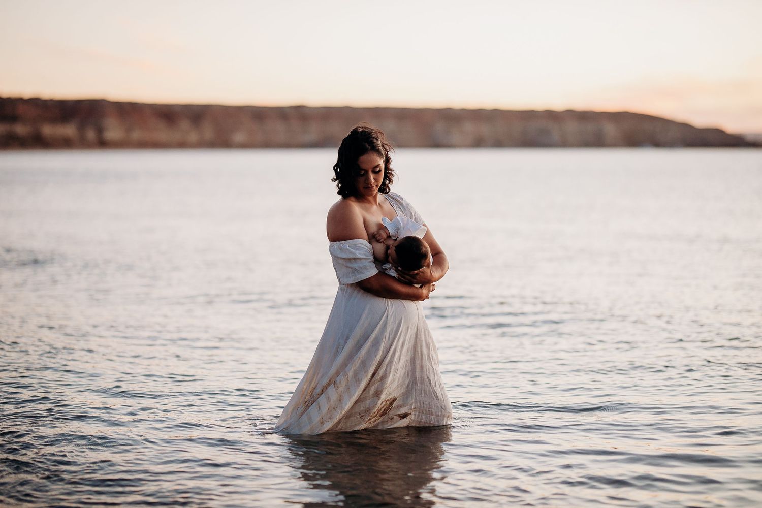 Mum standing in shallow ocean holding baby with coastal cliffs in background – Raised by Water Photography, Adelaide