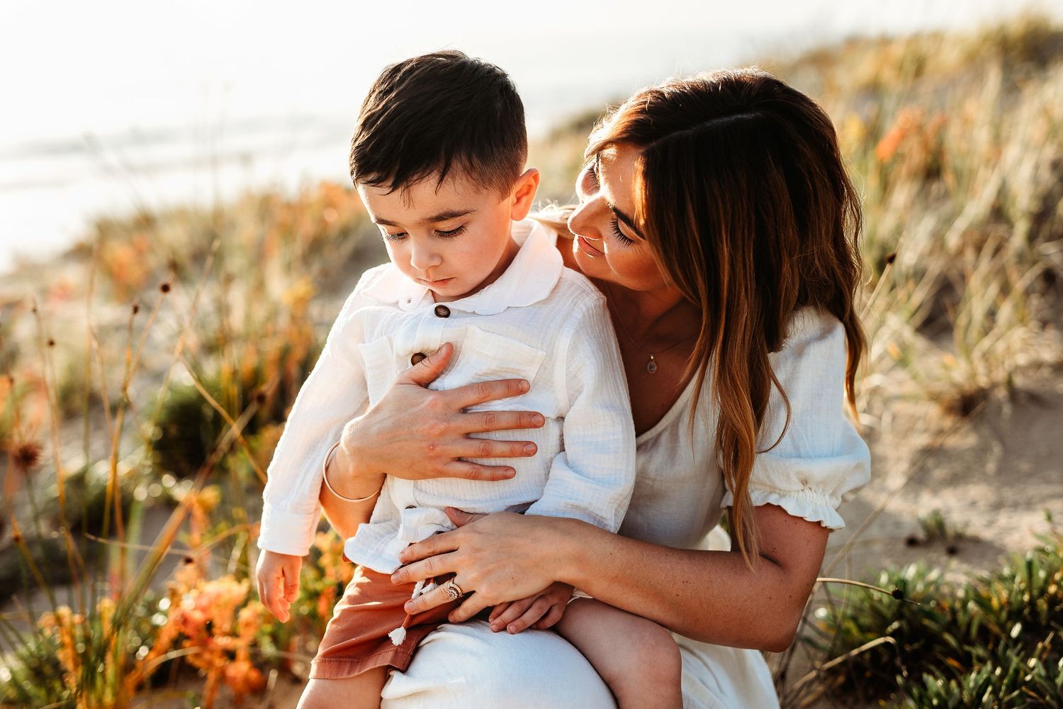 Mum looking lovingly at her child sitting on sand dunes surrounded by flowers.  Adelaide family photographer - Raised by Water Photography