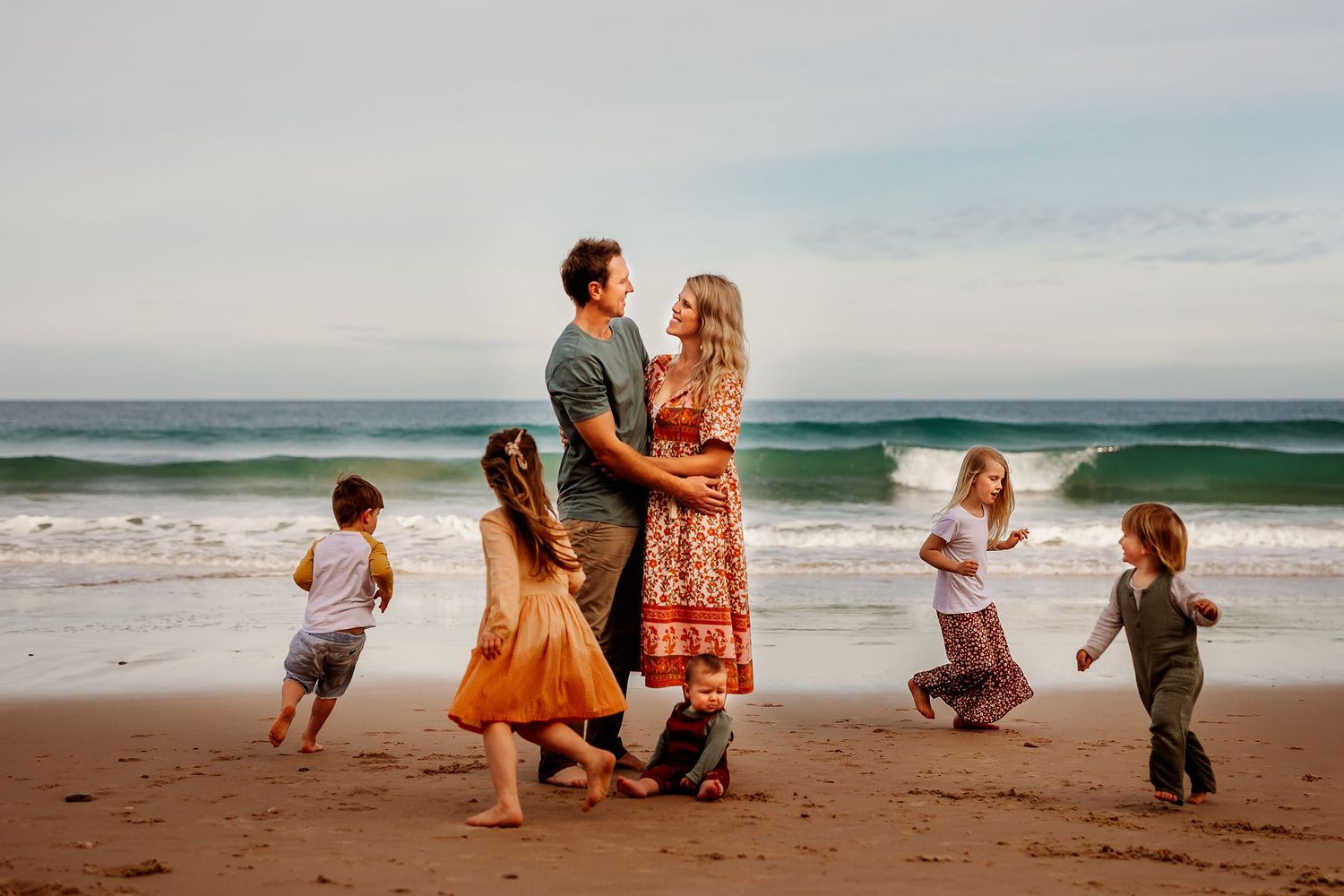 Mum and Dad embracing while children run around them playing.  Adelaide, South Australia family lifestyle photographer
