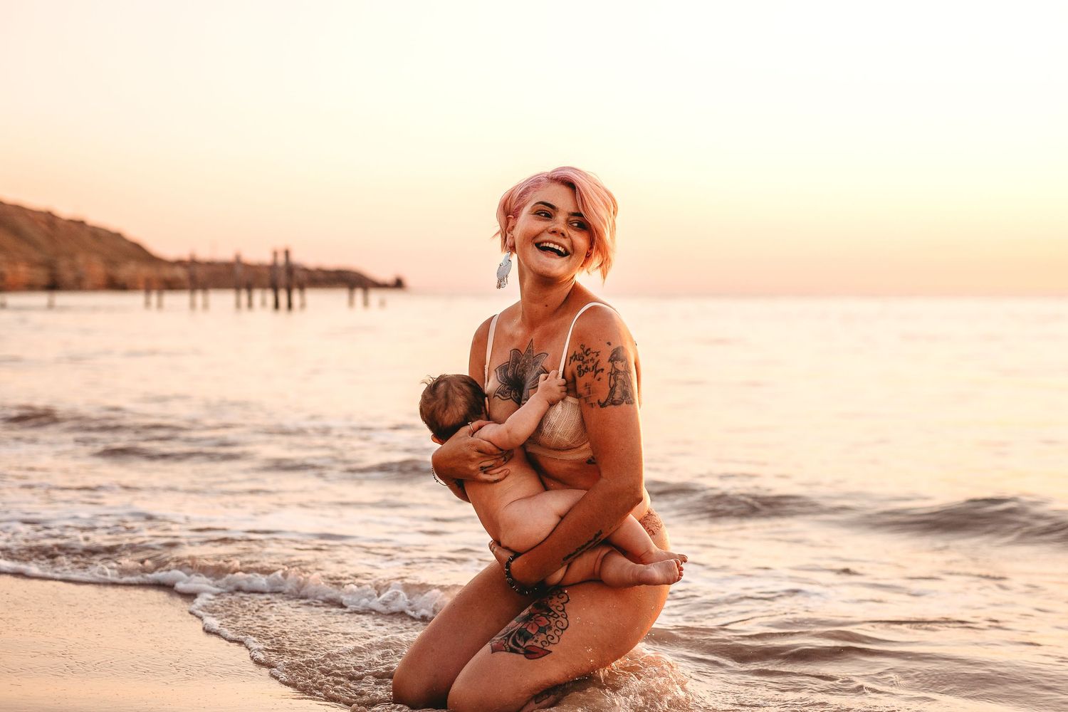 Mum sitting at the water’s edge laughing while holding baby – Raised by Water Photography, Adelaide