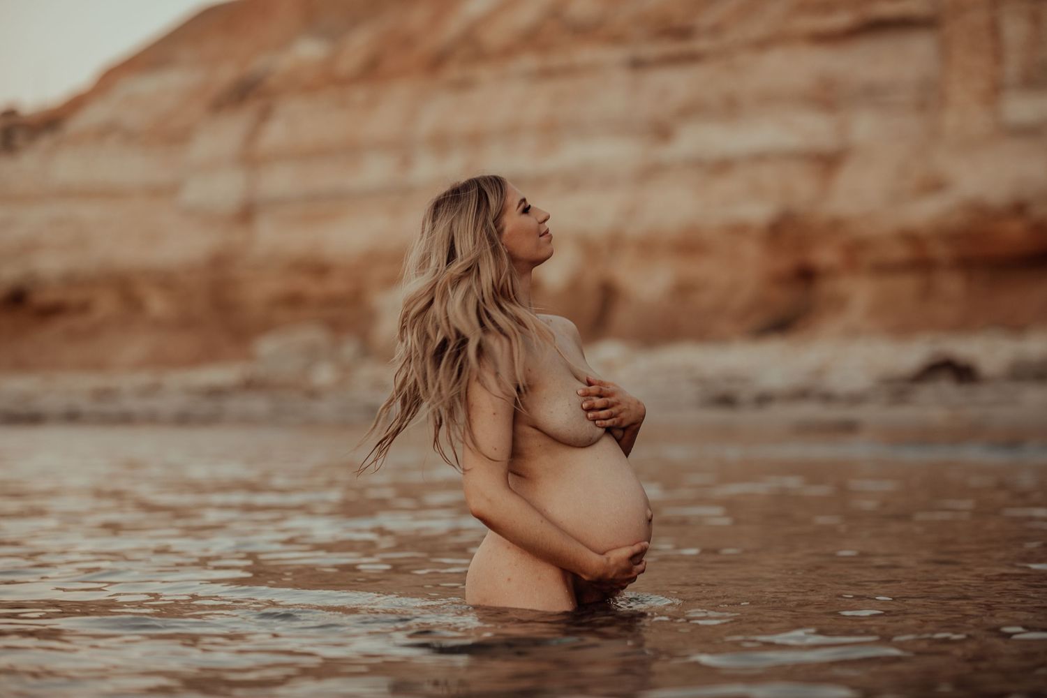 Pregnant woman standing in gentle waves at sunset, holding belly – Natural light session
