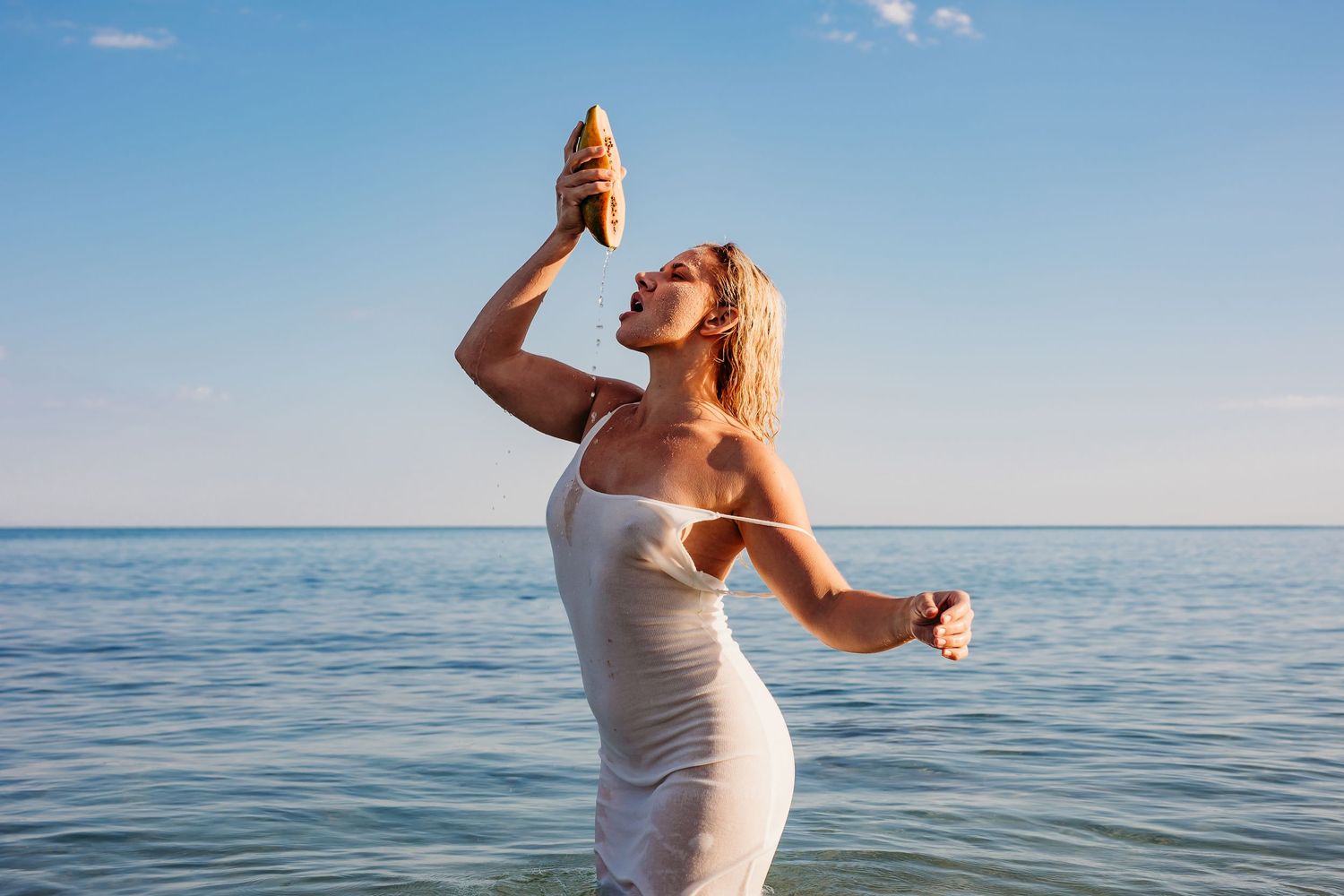 Lady standing in ocean wearing white dress squeezing fresh papaya juice into her mouth – Raised by Water Photography, Adelaide