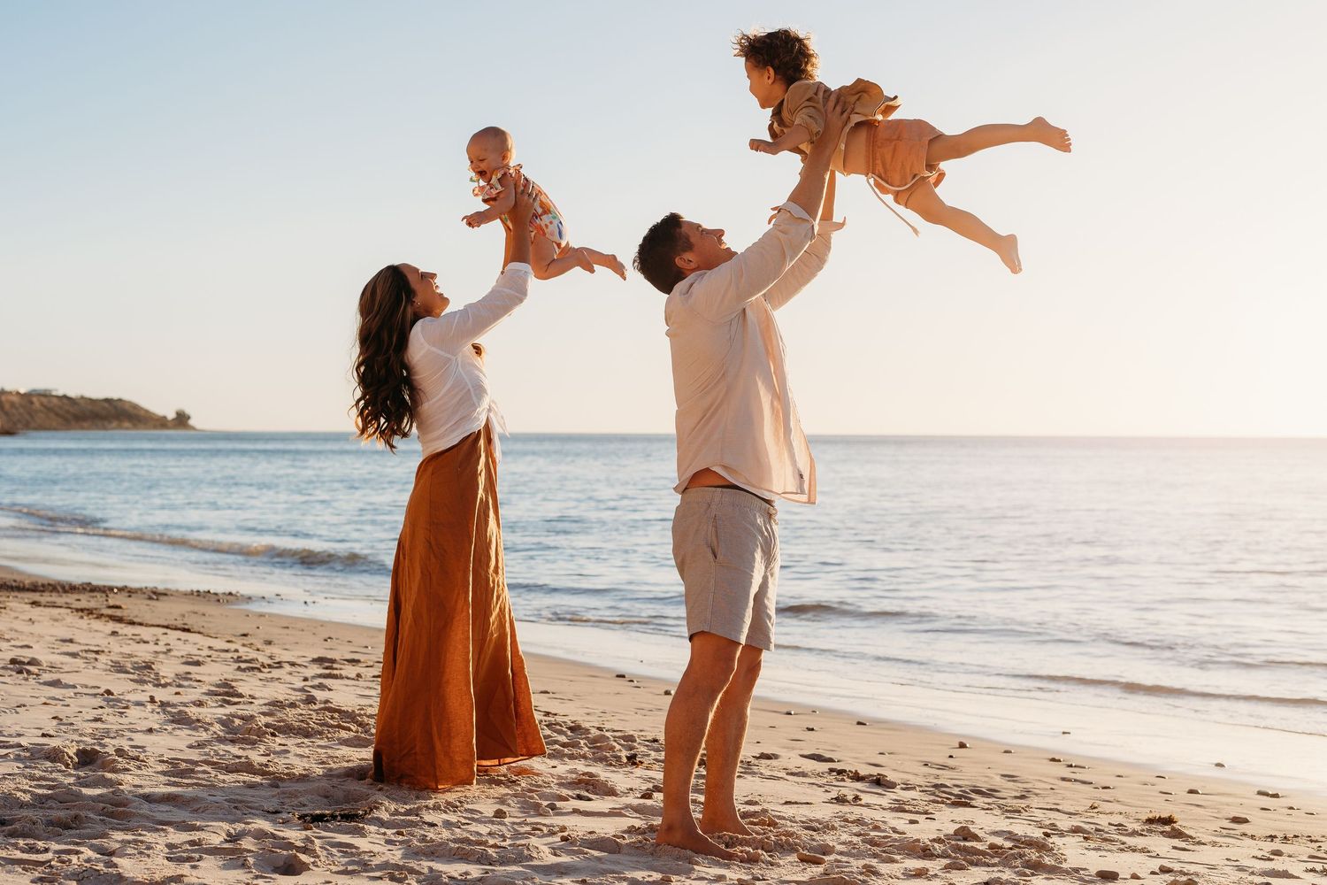 Mum and dad joyfully lifting their children high in the air at the beach – Raised by Water Photography, Adelaide
