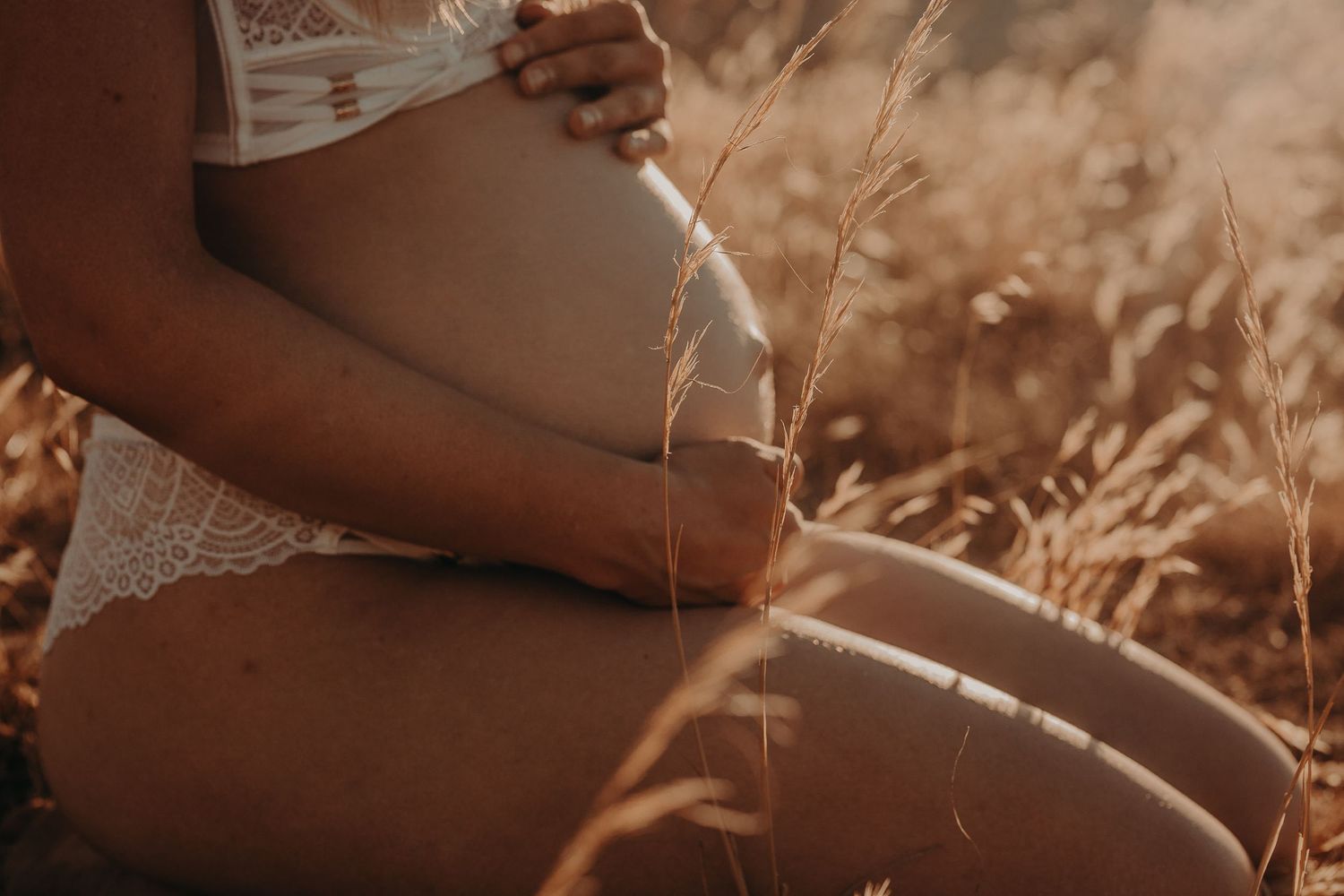 Close-up of baby bump while sitting in golden grass at sunset – Raised by Water Photography, Adelaide