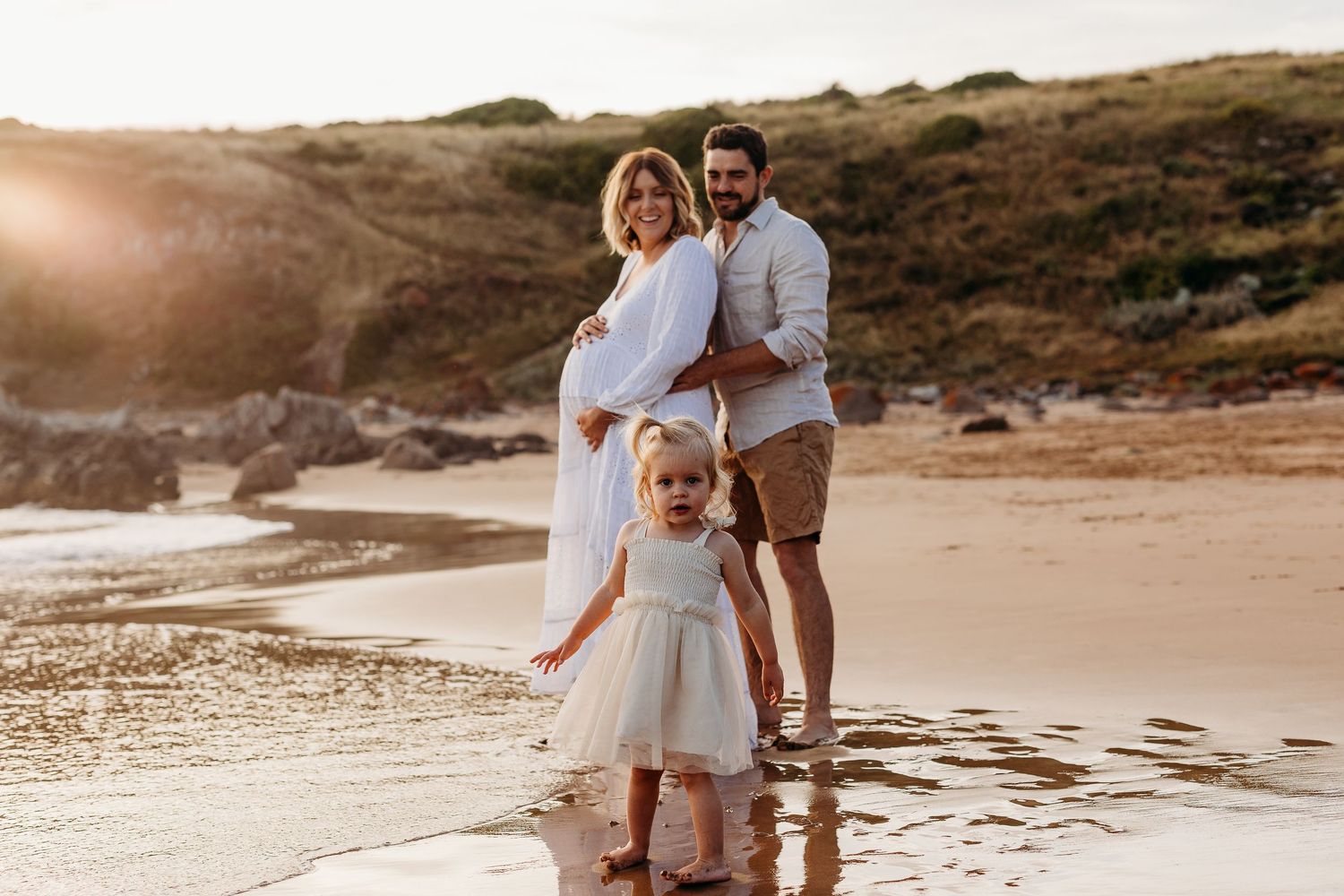 Pregnant mum with partner embracing her belly with daughter playing near them on a beach in Adelaide.