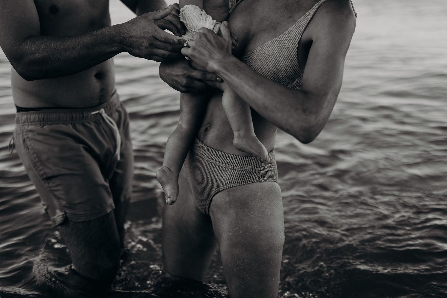 Close-up of mum, dad, and baby legs standing together in shallow ocean water – Raised by Water Photography, Adelaide