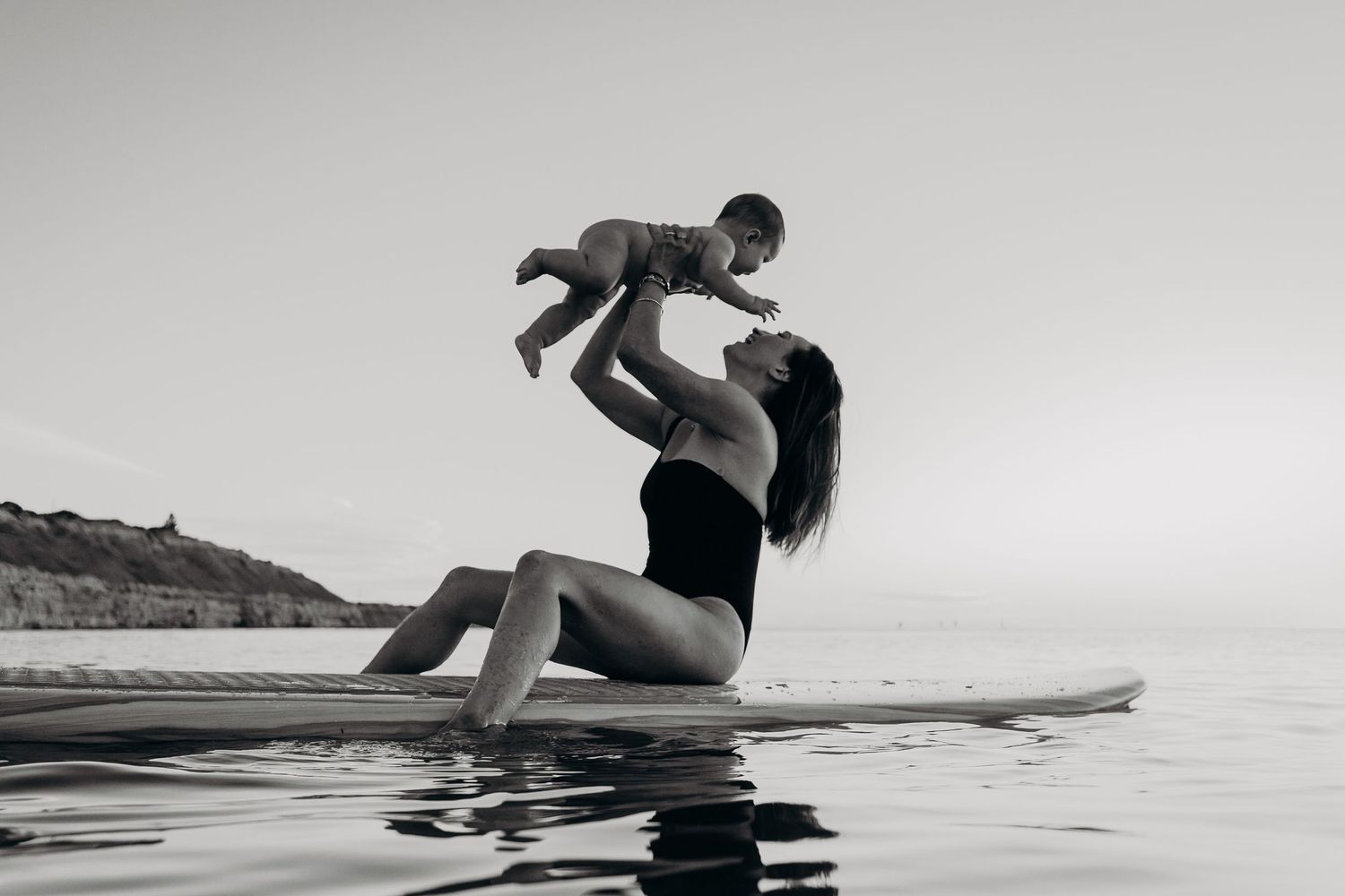 Candid photo of mother lifting baby while sitting on surfboard in the sea – South Australia family photography