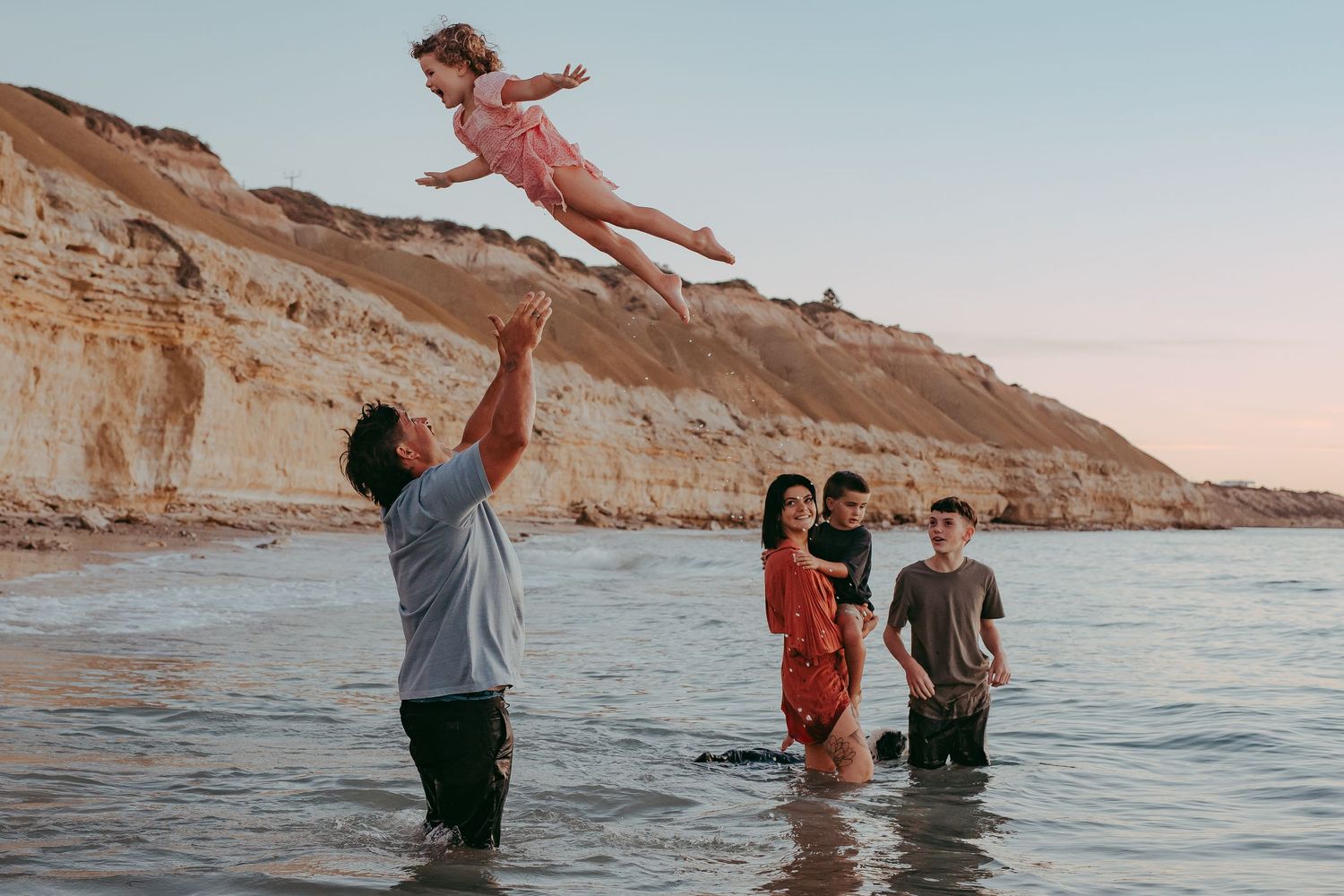 Family standing in shallow water with dad playfully throwing daughter in the air – Adelaide beach photography