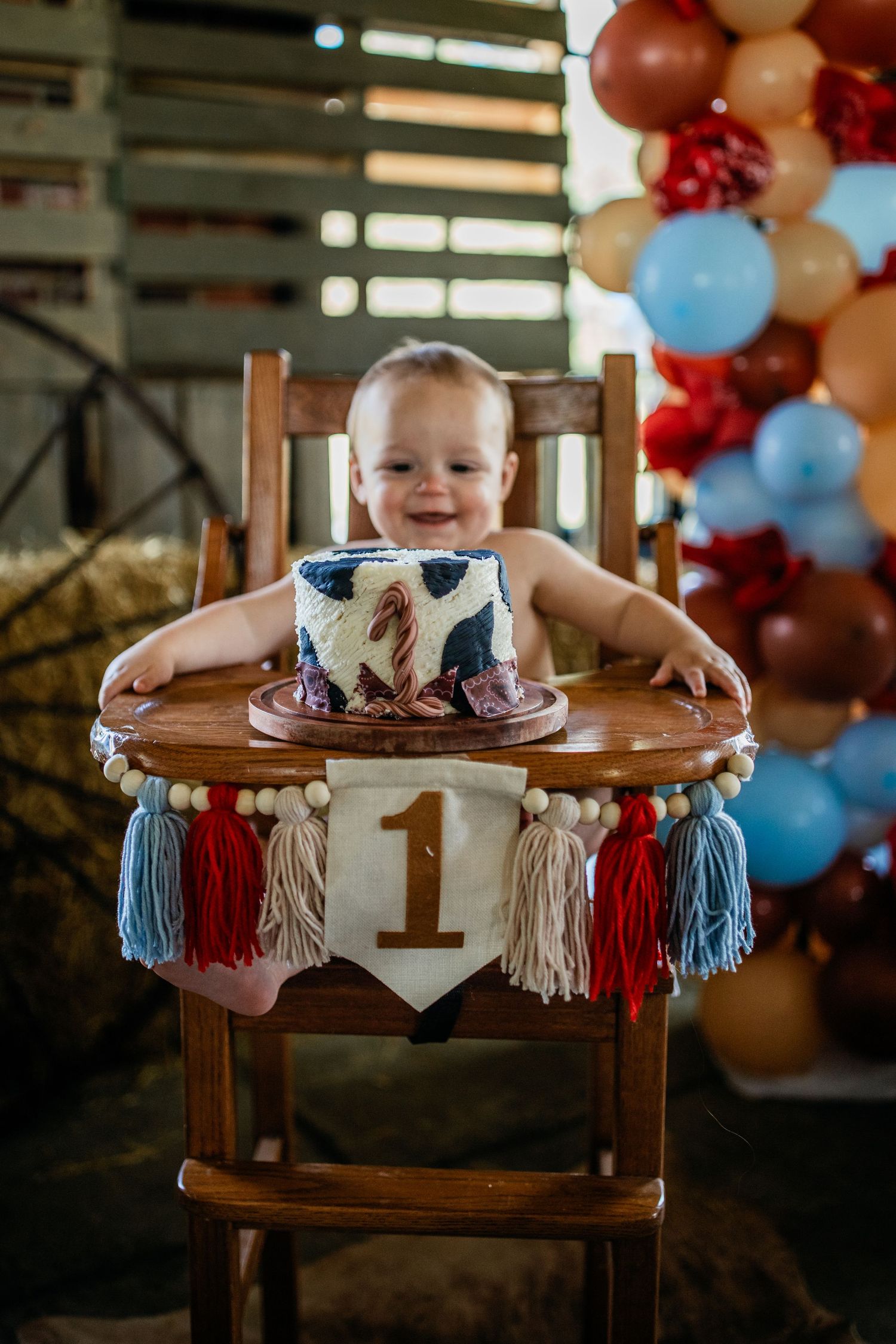 Baby sits in decorated highchair with first birthday cake and balloon arch backdrop.