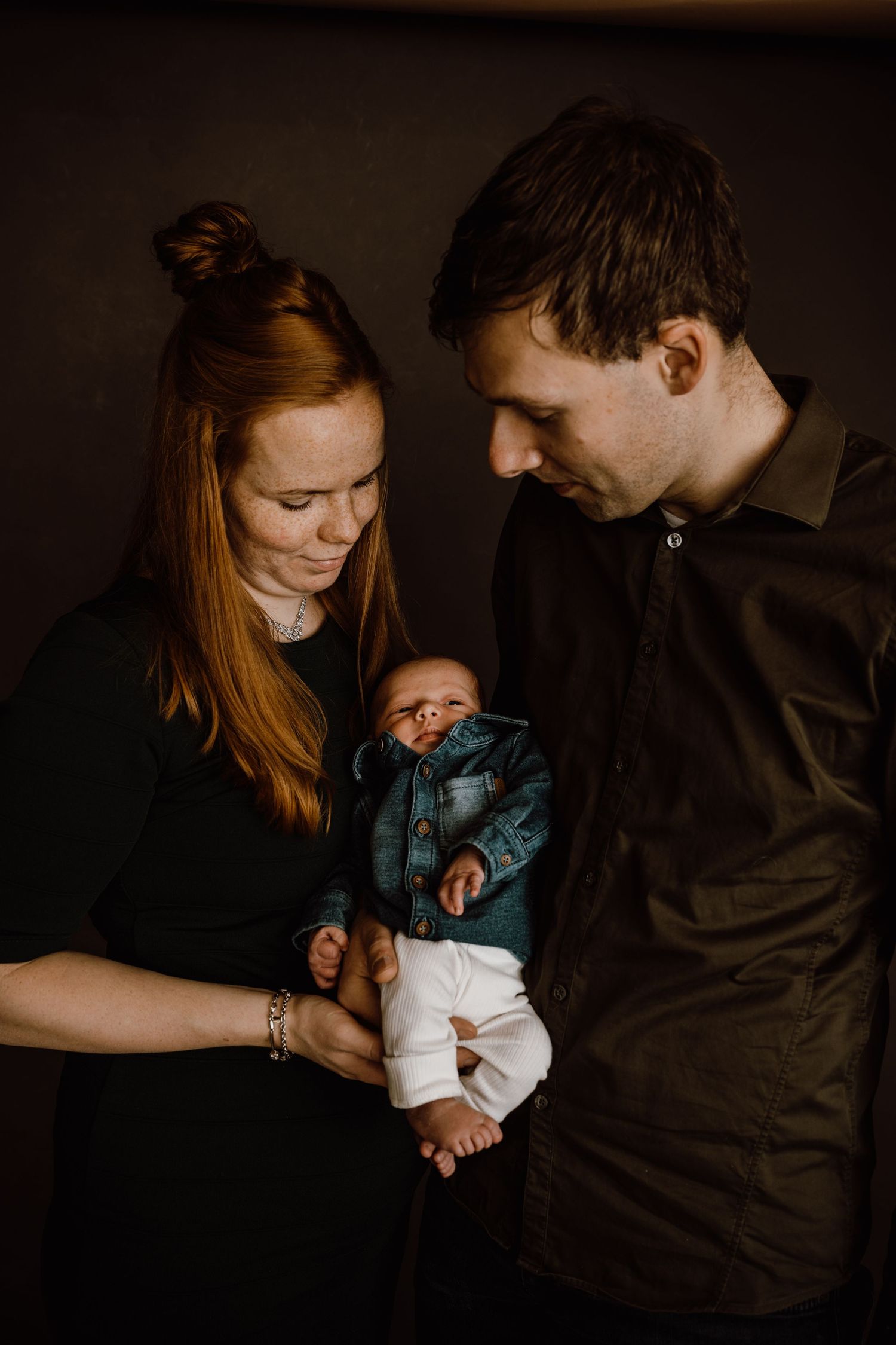 A series of intimate dark-toned photographs showing tender moments between parents and their newborn baby in dim lighting.