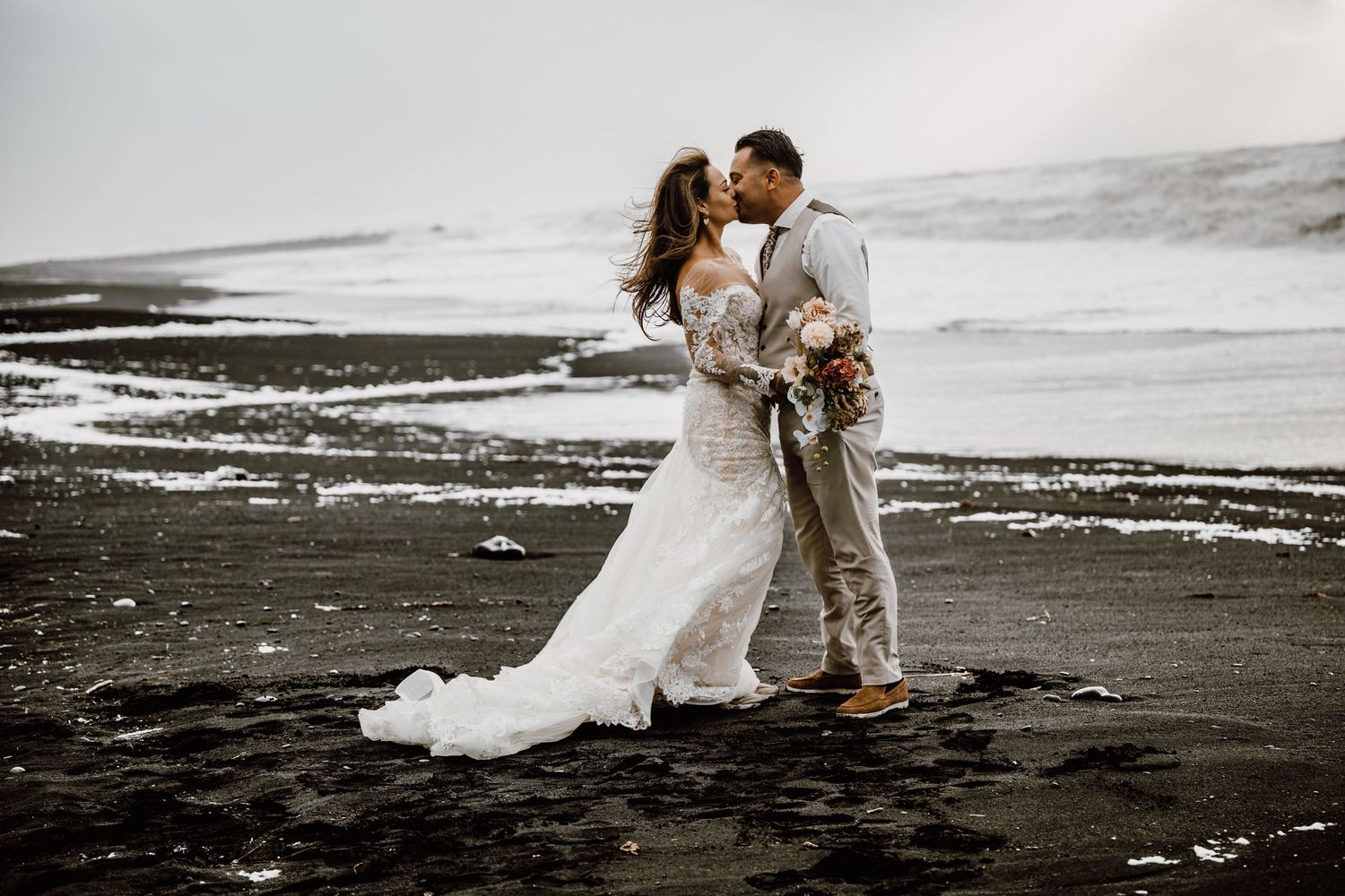 A couple shares an intimate moment on a dramatic black sand beach during their wedding photoshoot.